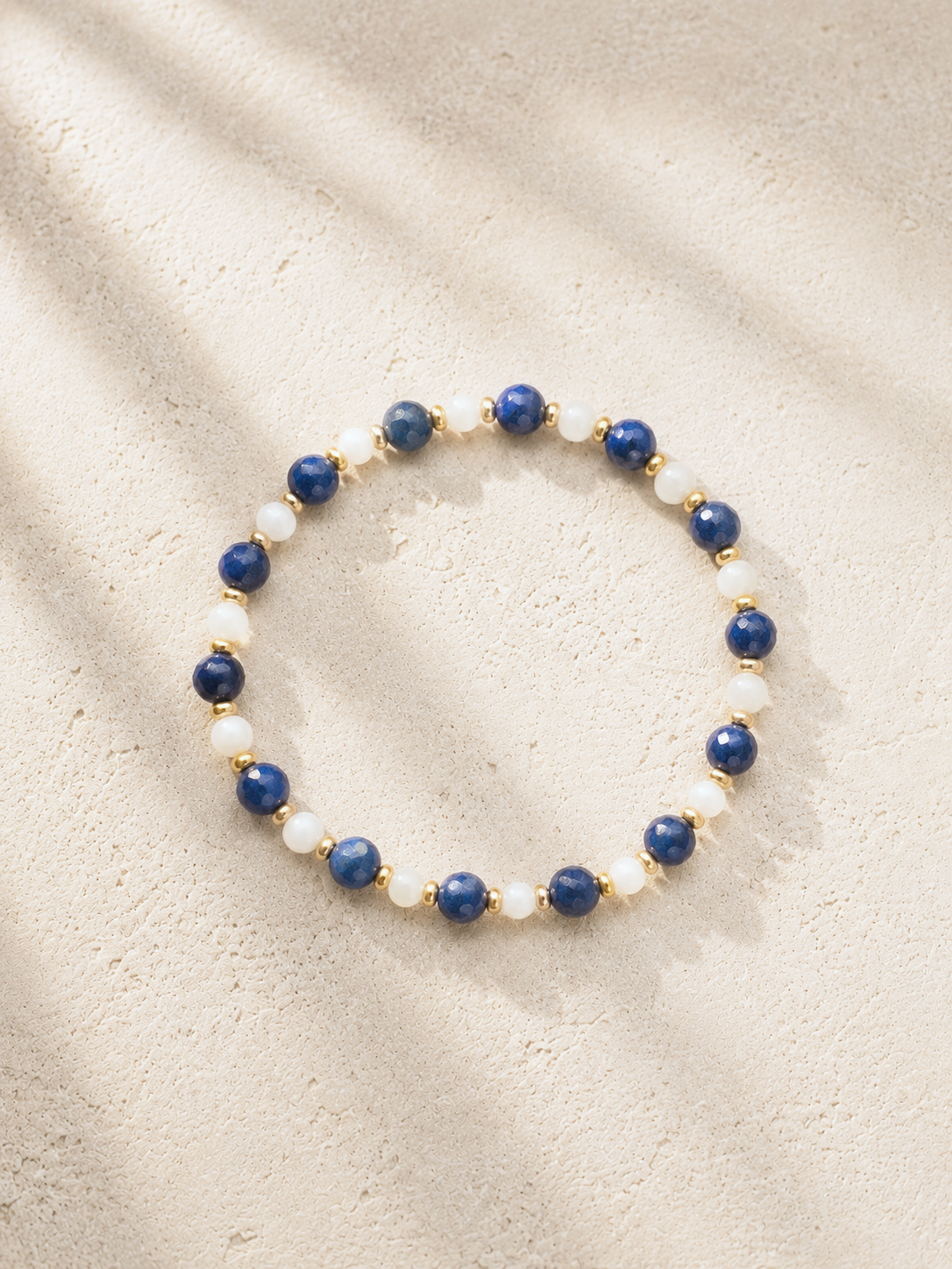 A beaded bracelet with alternating dark blue and white beads, accented with small gold beads, resting on a sandy surface with shadow patterns.