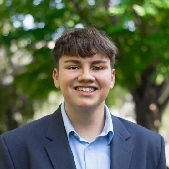 A young man with brown hair, wearing a blue blazer and a light blue shirt, smiling outdoors with green trees in the background.