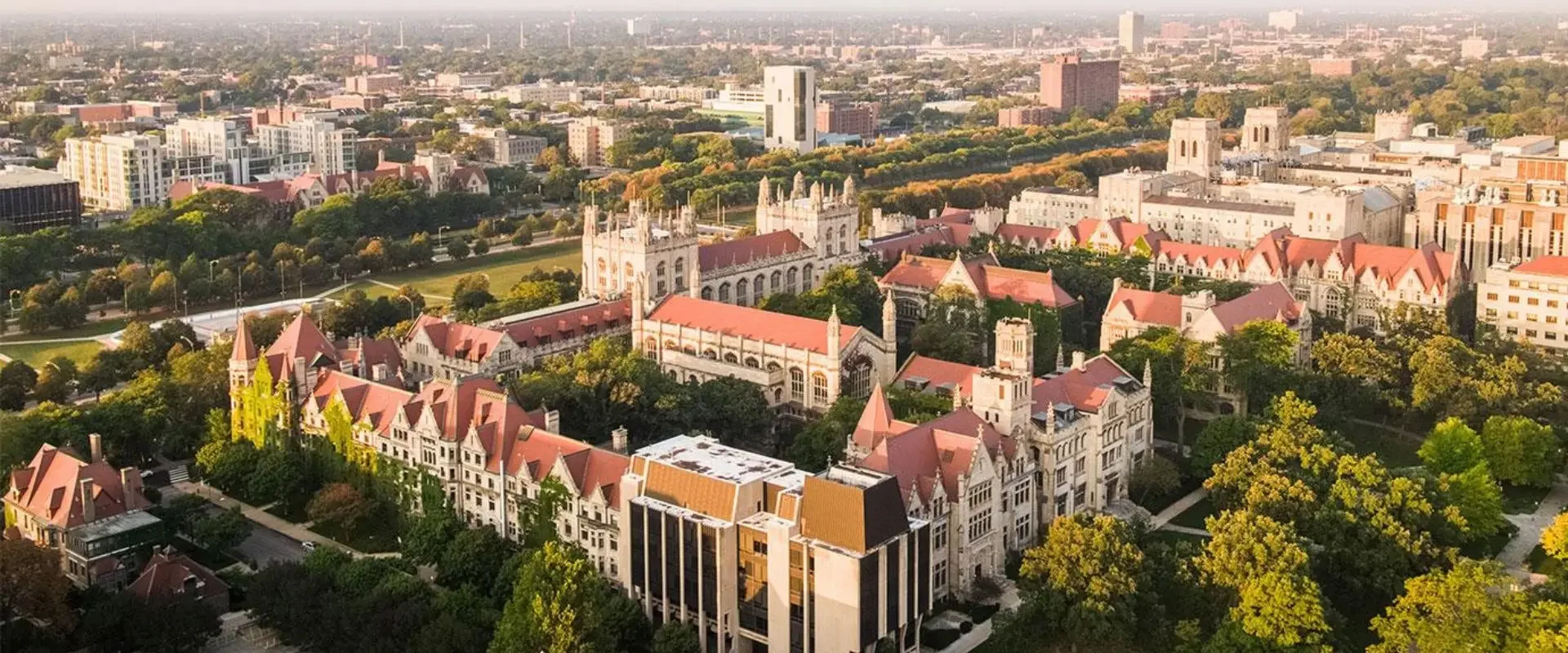 Aerial view of a historic university campus with gothic-style buildings featuring red-tiled roofs surrounded by green trees, cityscape in the background.