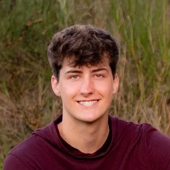 A young man with brown hair and a bright smile, outdoors in front of tall grass.
