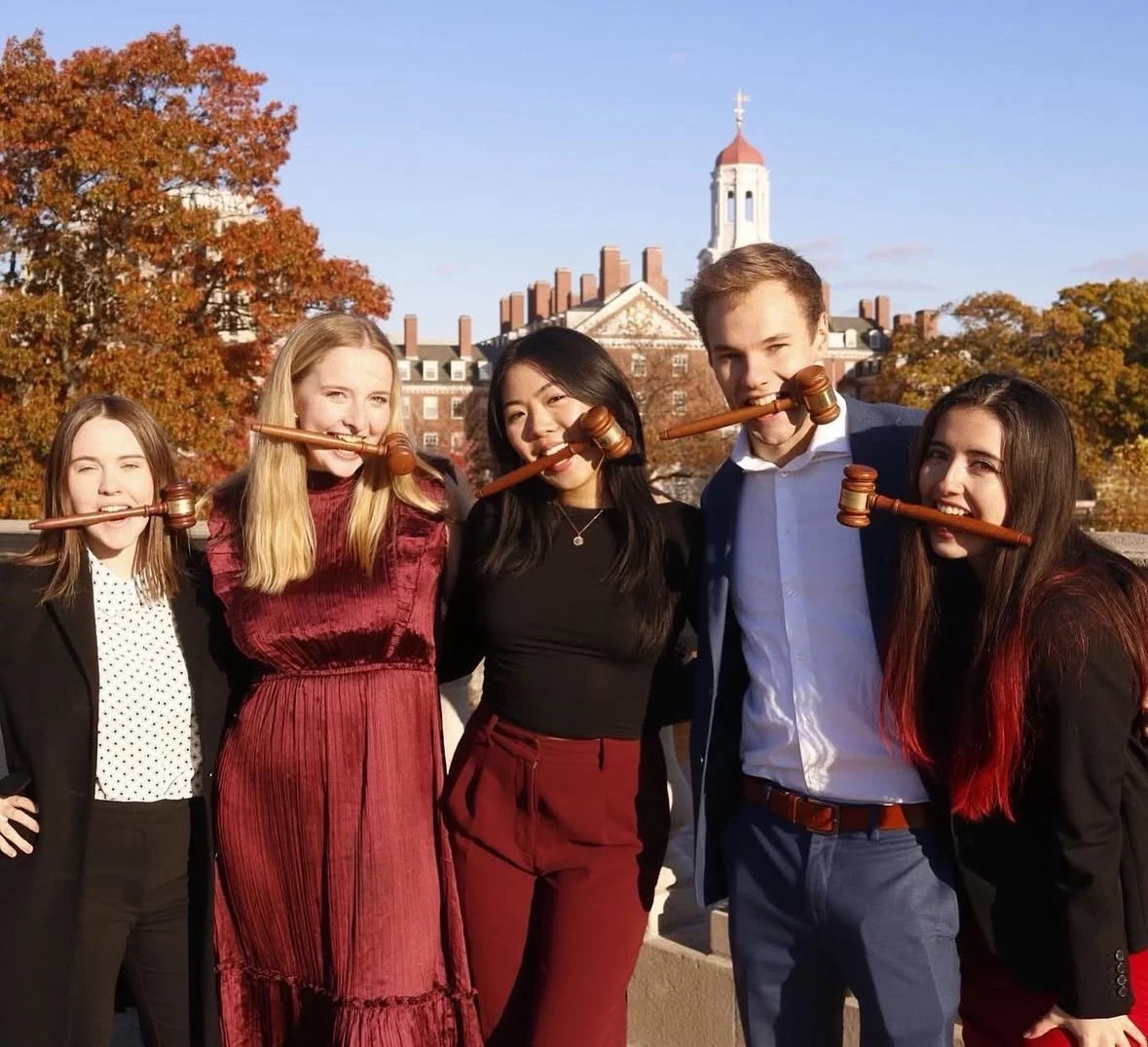 Five young adults standing outdoors during daytime, holding wooden gavel props in their mouths, with historical buildings and autumn trees in the background.