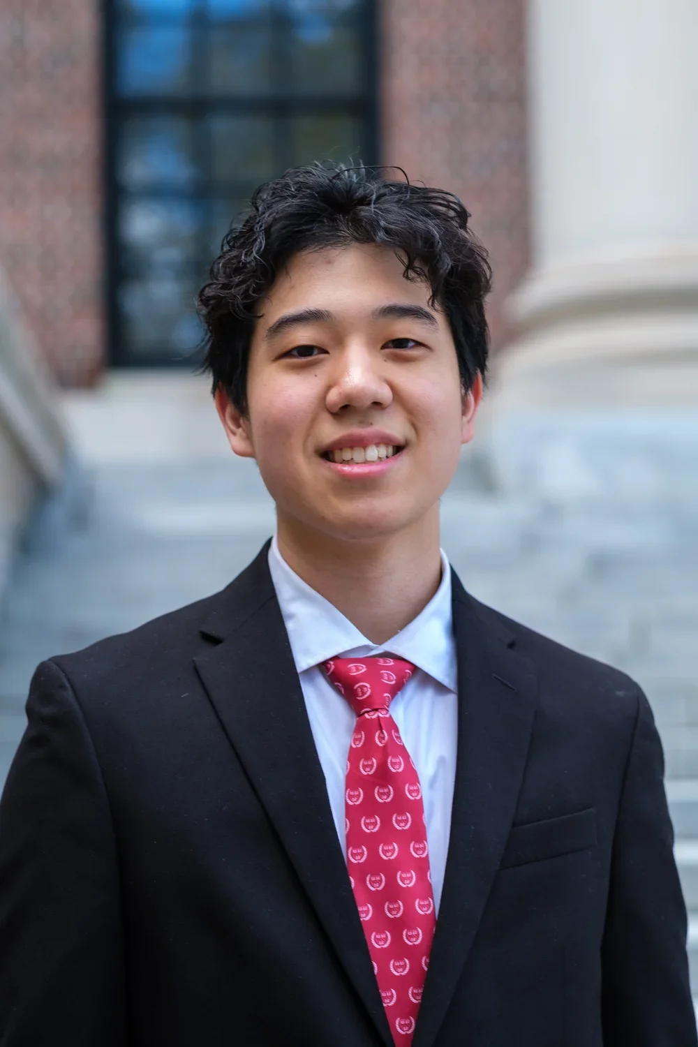 Portrait of a young man with black curly hair wearing a black suit, white shirt, and red tie, standing outdoors on stairs in front of a brick and glass building.