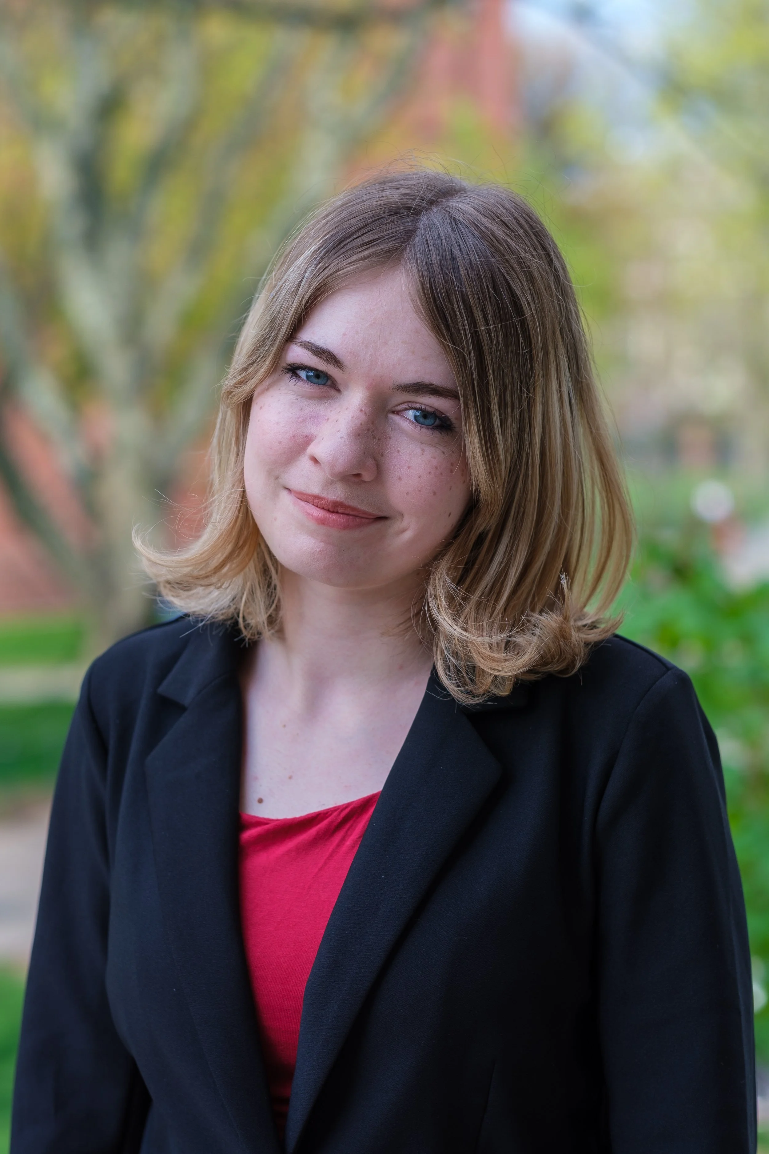 A young woman with shoulder-length light brown hair and blue eyes, smiling slightly, outdoors with blurred trees in the background, wearing a black blazer over a red top.