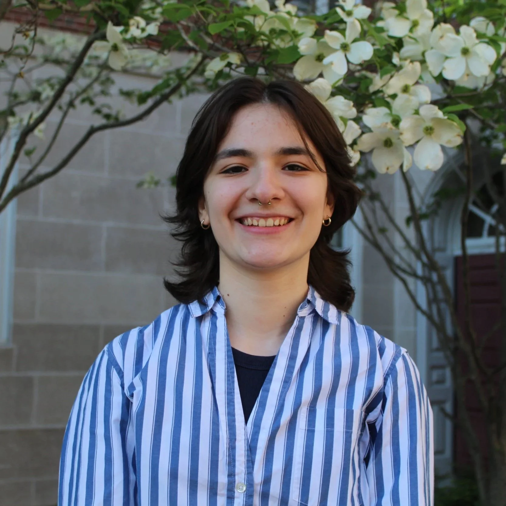 A young woman with short dark hair and a septum piercing smiling outdoors, standing in front of a blooming dogwood tree with white flowers.