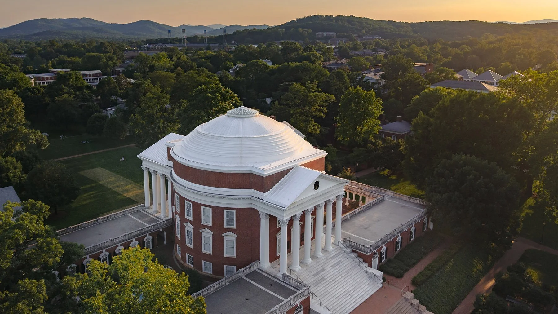 Aerial view of a historic domed building with white columns, red brick walls, and a white roof, surrounded by lush green trees in a hilly landscape at sunset.