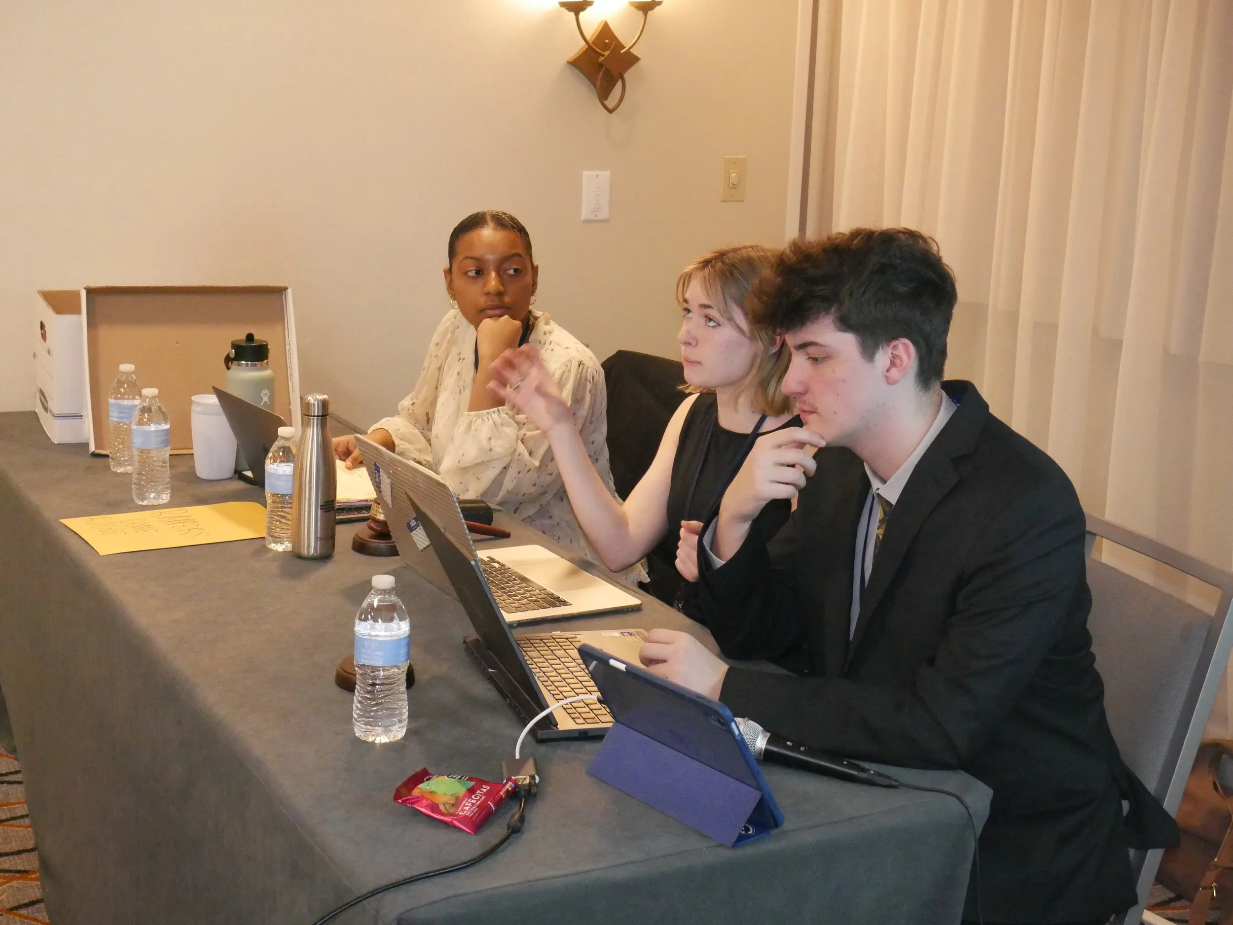 Three people sitting at a conference table with laptops, water bottles, and snacks, engaged in a discussion in a well-lit room with cream walls and a curtain.