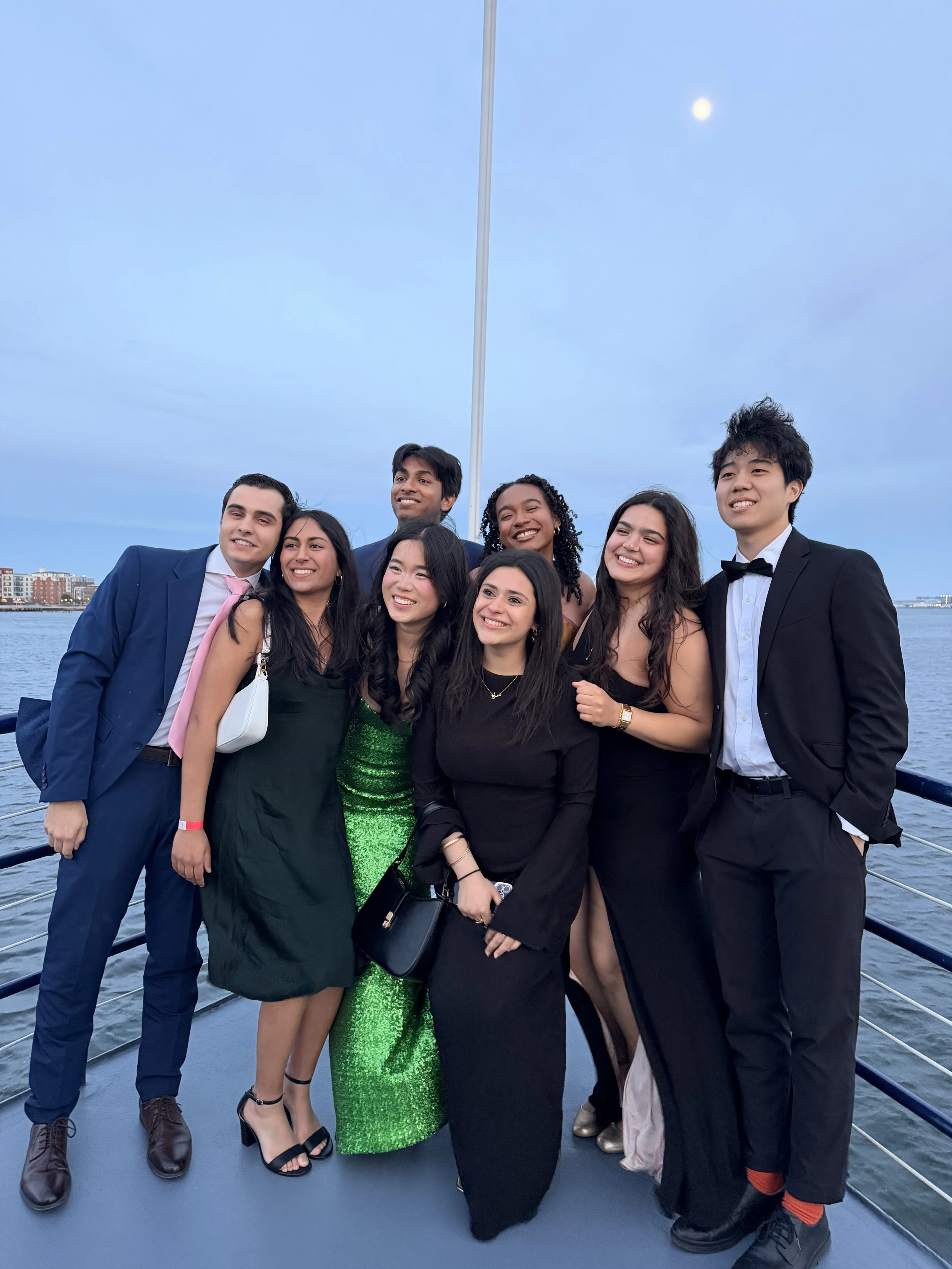 Group of nine young adults dressed in formal attire, smiling and posing on a boat with a city skyline and water in the background, during dusk or evening.
