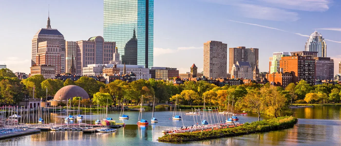 City skyline with modern skyscrapers, green trees, and sailboats on a body of water in foreground.