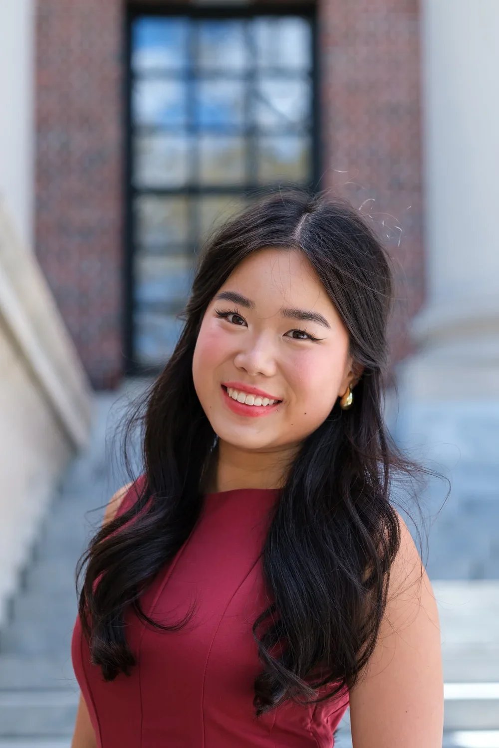 A young woman with long black hair, smiling, wearing a sleeveless maroon dress and pearl earrings, outdoors near a building with stairs in the background.