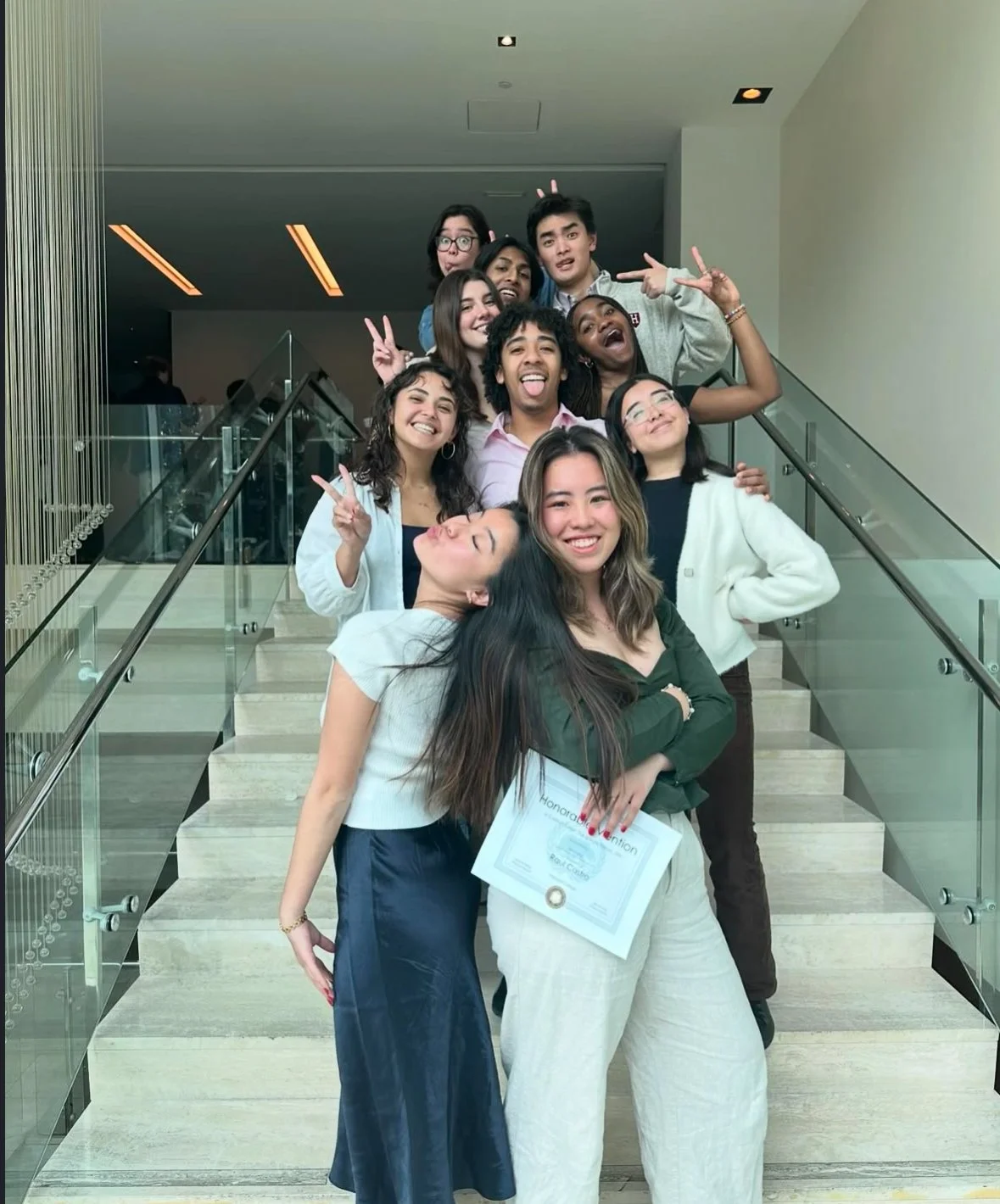 Group of young adults posing on a staircase indoors, some making peace signs, one holding a certificate, all smiling and having fun.