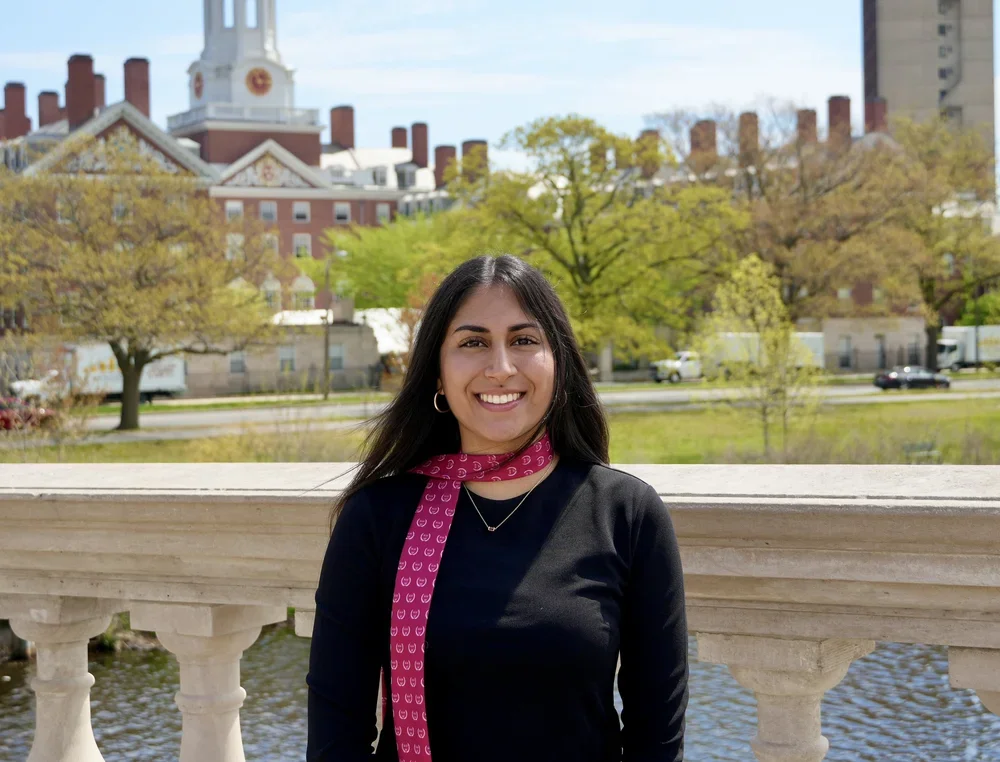 A woman standing outdoors on a stone bridge with a cityscape and trees in the background. She is smiling, wearing a black top, hoop earrings, a pink patterned scarf, and a small gold necklace.