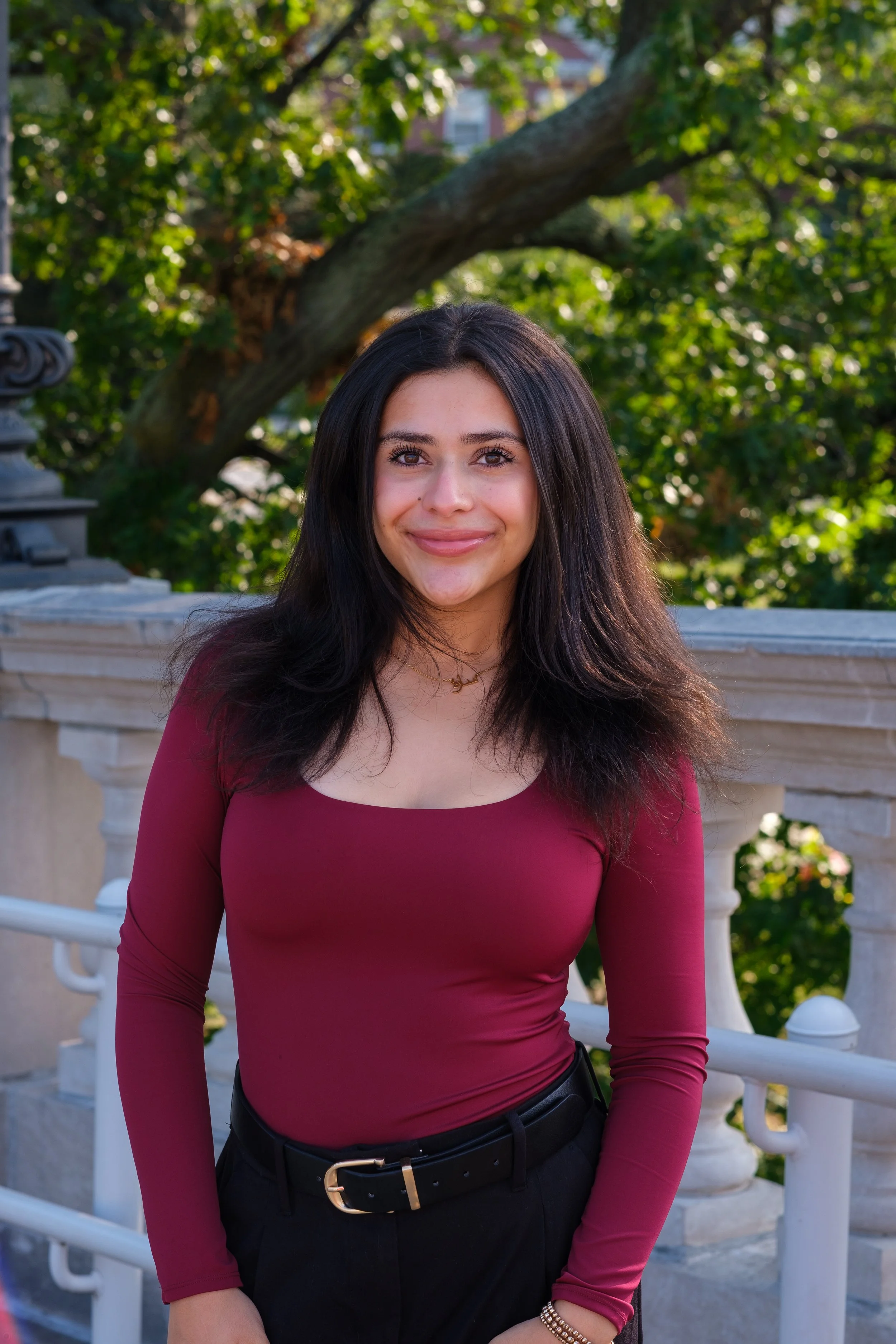 A young woman with long dark hair smiling outdoors near a stone balustrade and greenery.