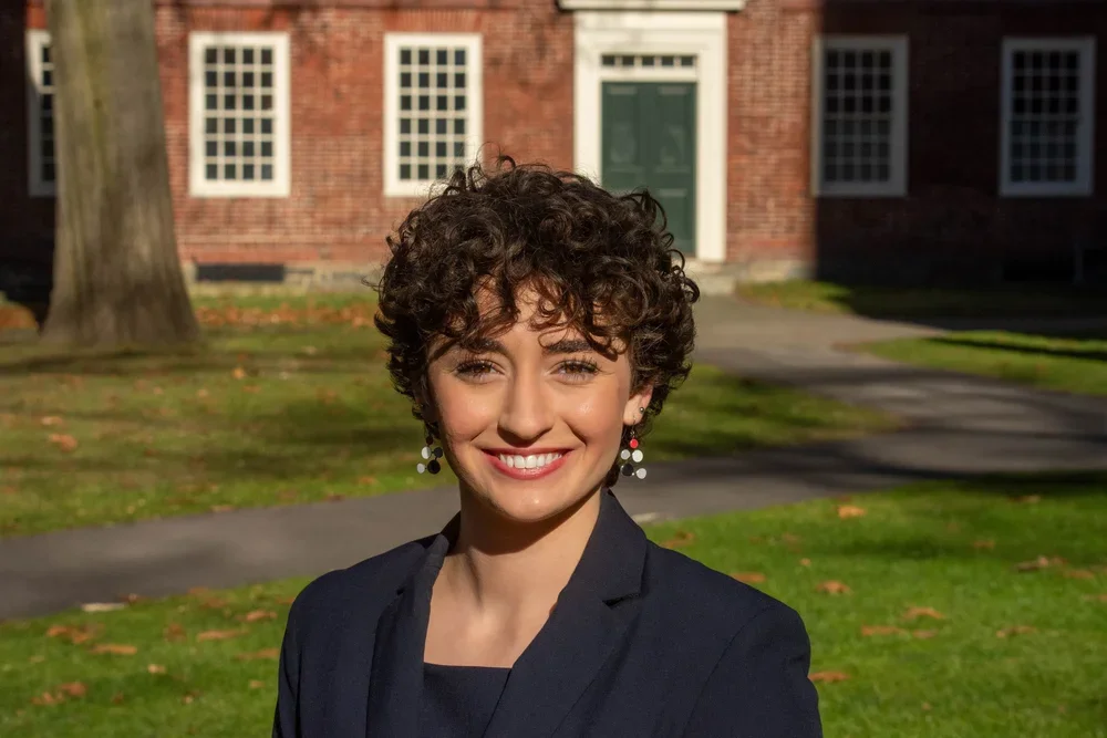 A woman with short, curly dark hair smiling outdoors in front of a brick building, wearing a black blazer and earrings.