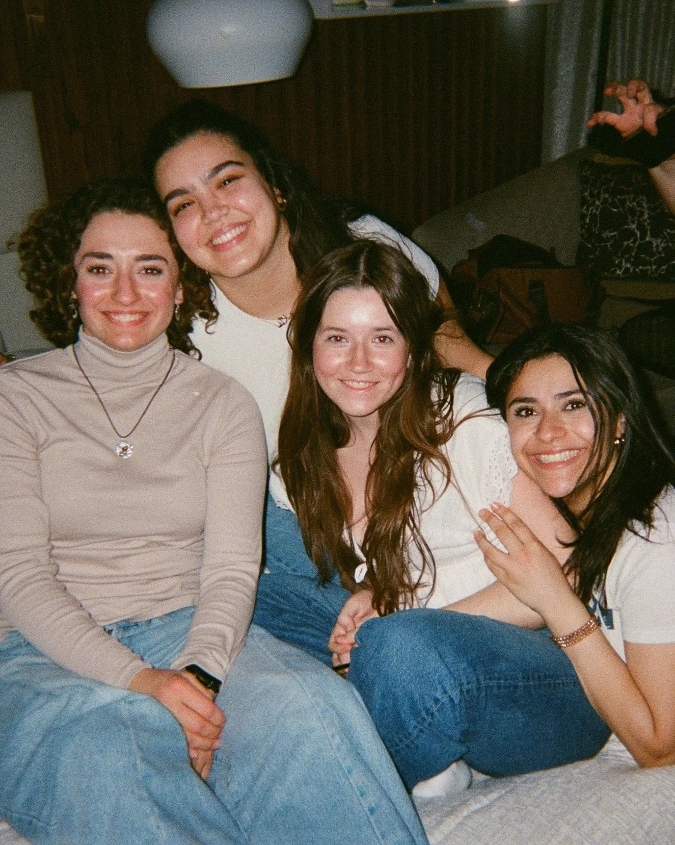A group of five women sitting closely together and smiling indoors with a brown wall and a patterned blanket in the background.