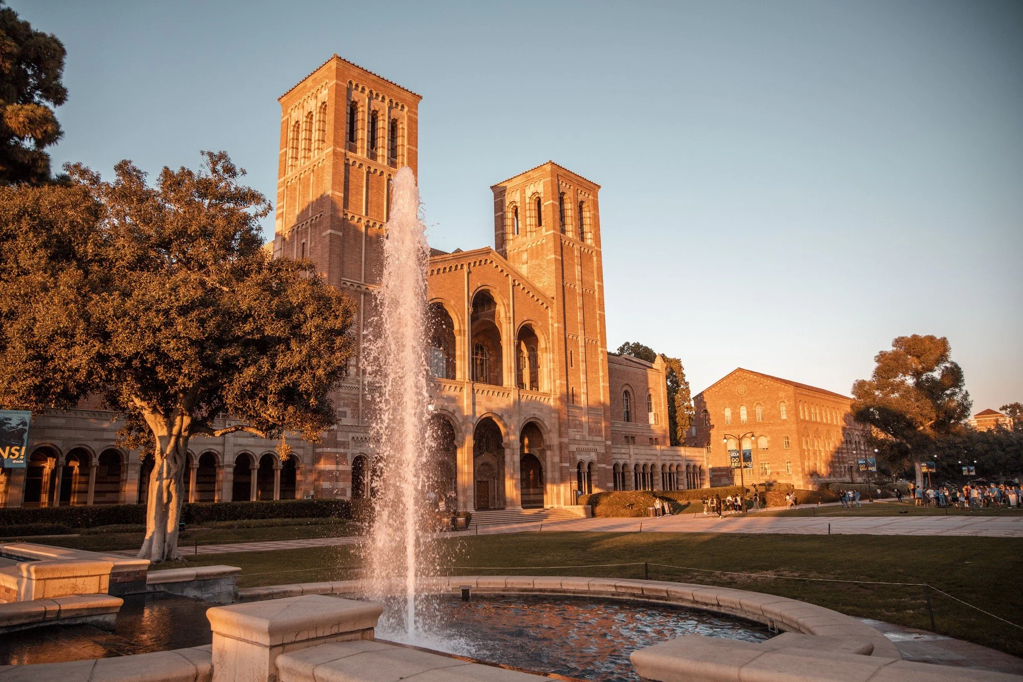 UCLA campus building with twin towers, a fountain in the foreground, trees, and people walking on campus during daylight.