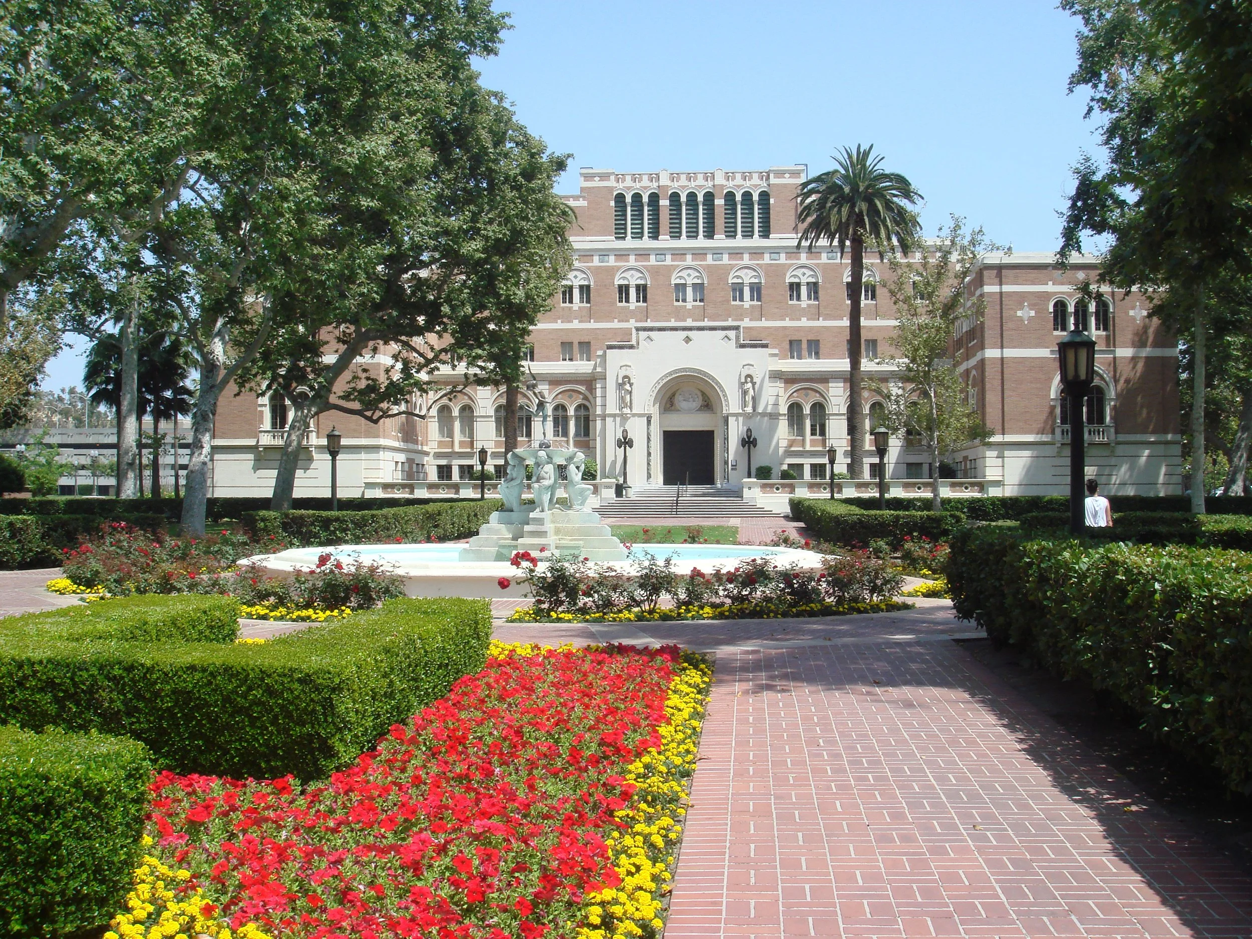 A historic brick building with arched windows surrounded by lush trees and a colorful flower bed in the foreground.