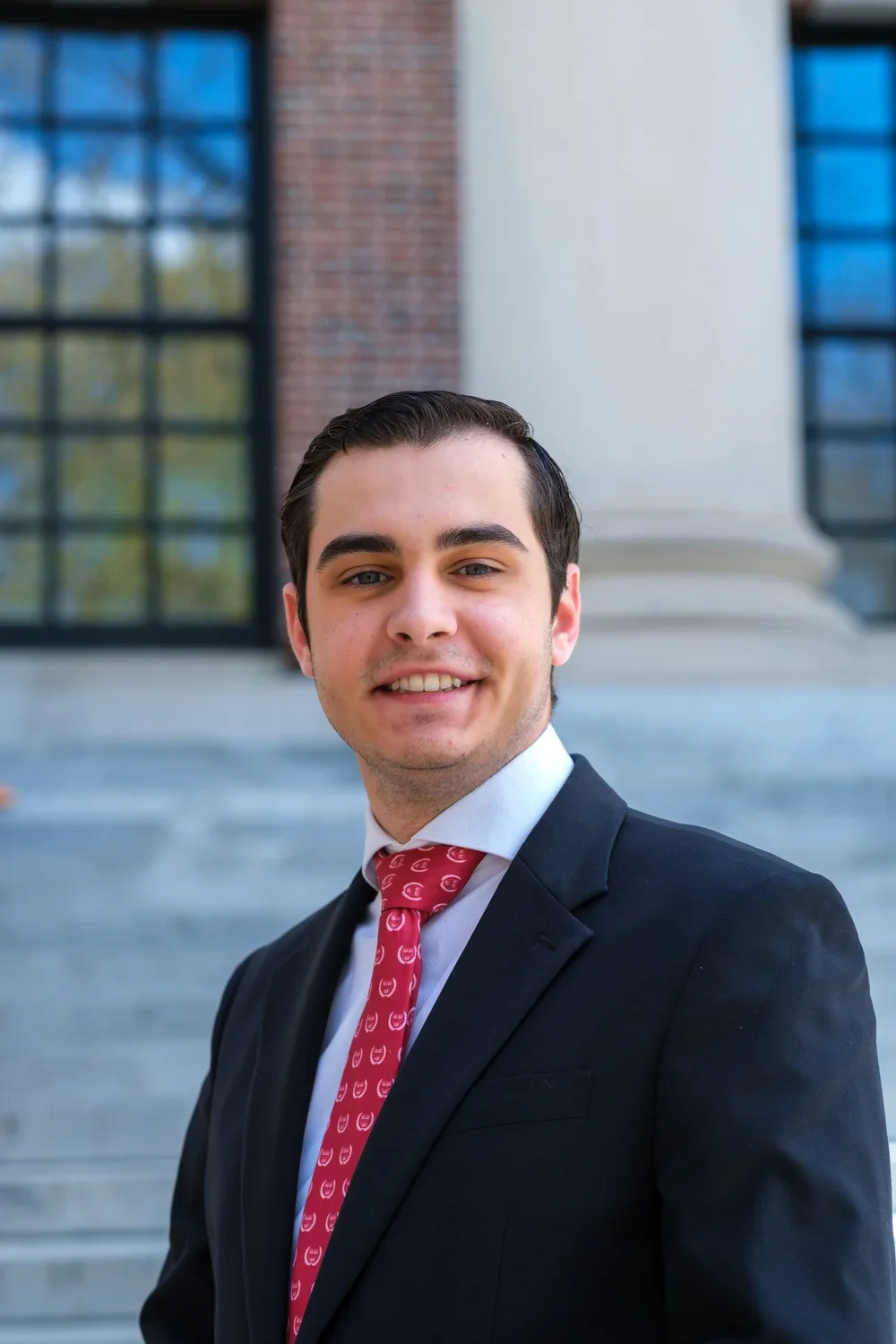 A young man wearing a dark suit, light shirt, and red patterned tie, standing outside a building with stone stairs and large windows.