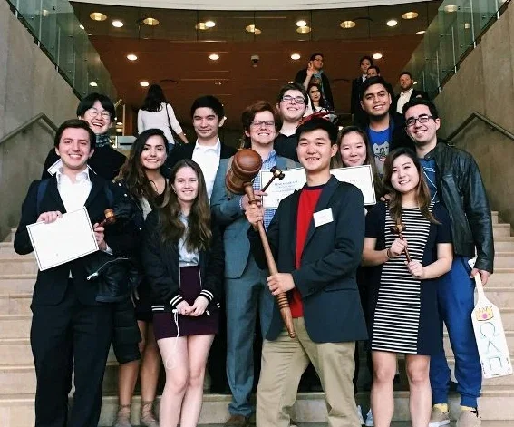 Group of young people smiling and posing on a staircase at an indoor event, some holding items like a gavel, a clipboard, and a sign.