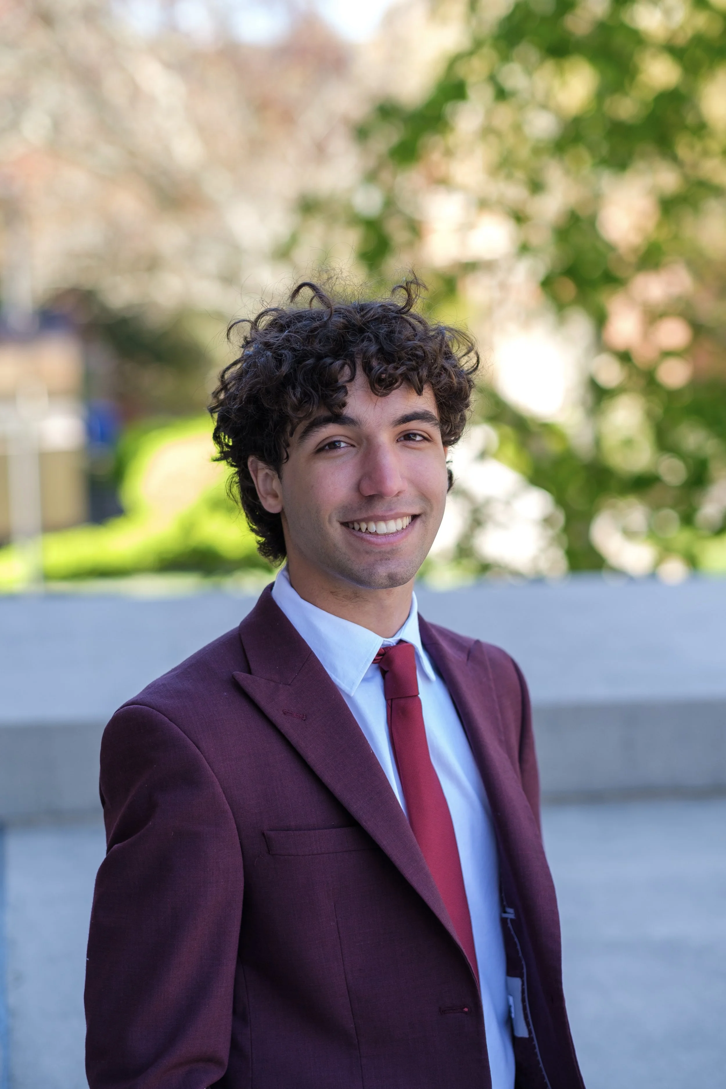 A young man in a maroon suit and red tie smiling outdoors with green trees and a bright background.