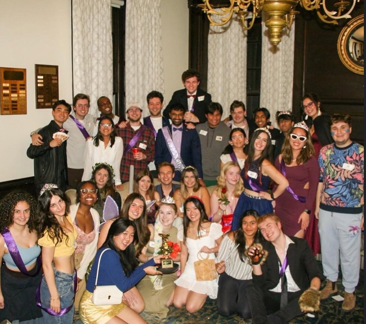 Group of young adults celebrating at a party or event, dressed in costumes and casual attire, with some wearing sashes and tiaras, in an elegant room with chandelier and decorative wall mirrors.