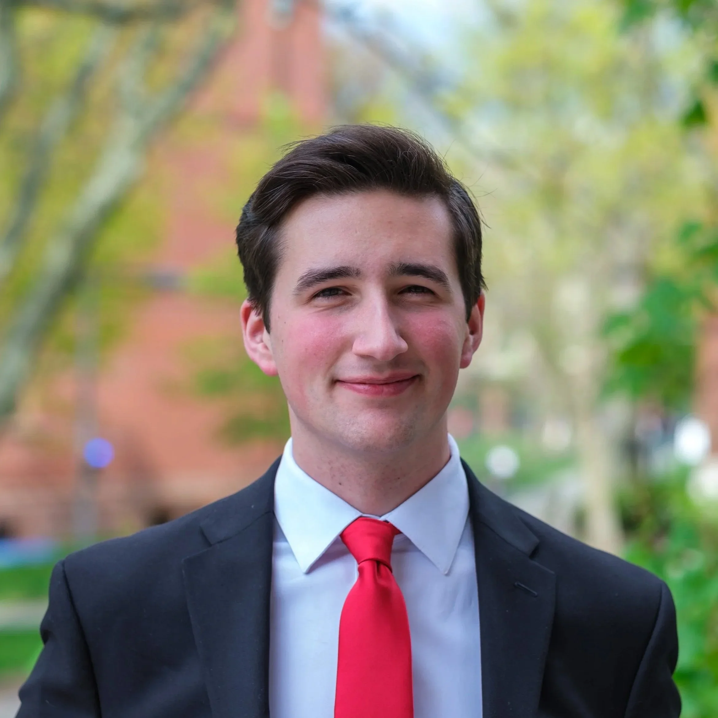 A young man smiling, dressed in a formal suit with a red tie, outdoors with blurred green and autumn-colored trees in the background.