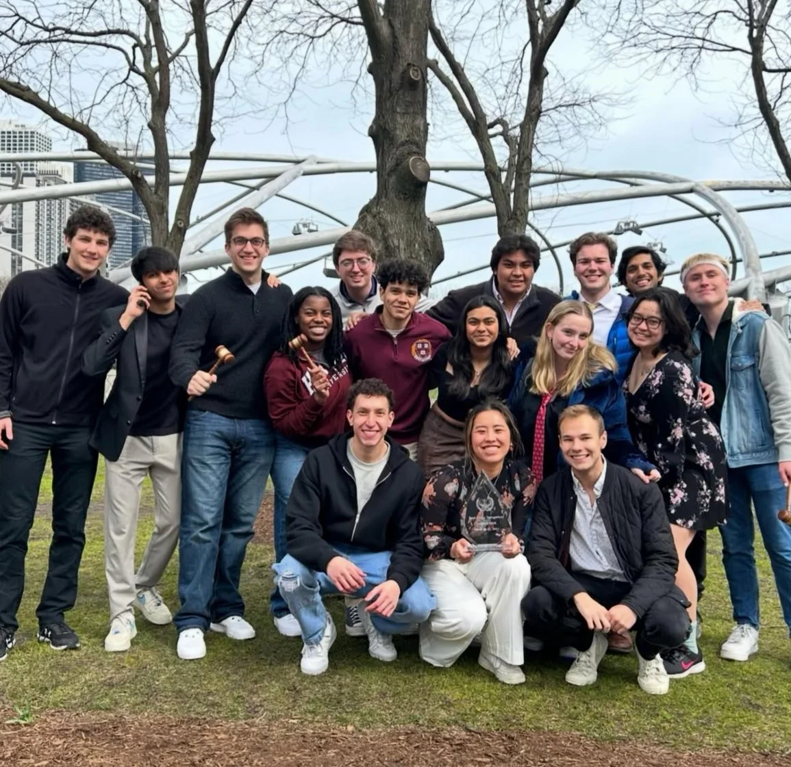 Group of young adults posing outdoors in front of a large tree and a modern, circular metallic structure, smiling for the camera.
