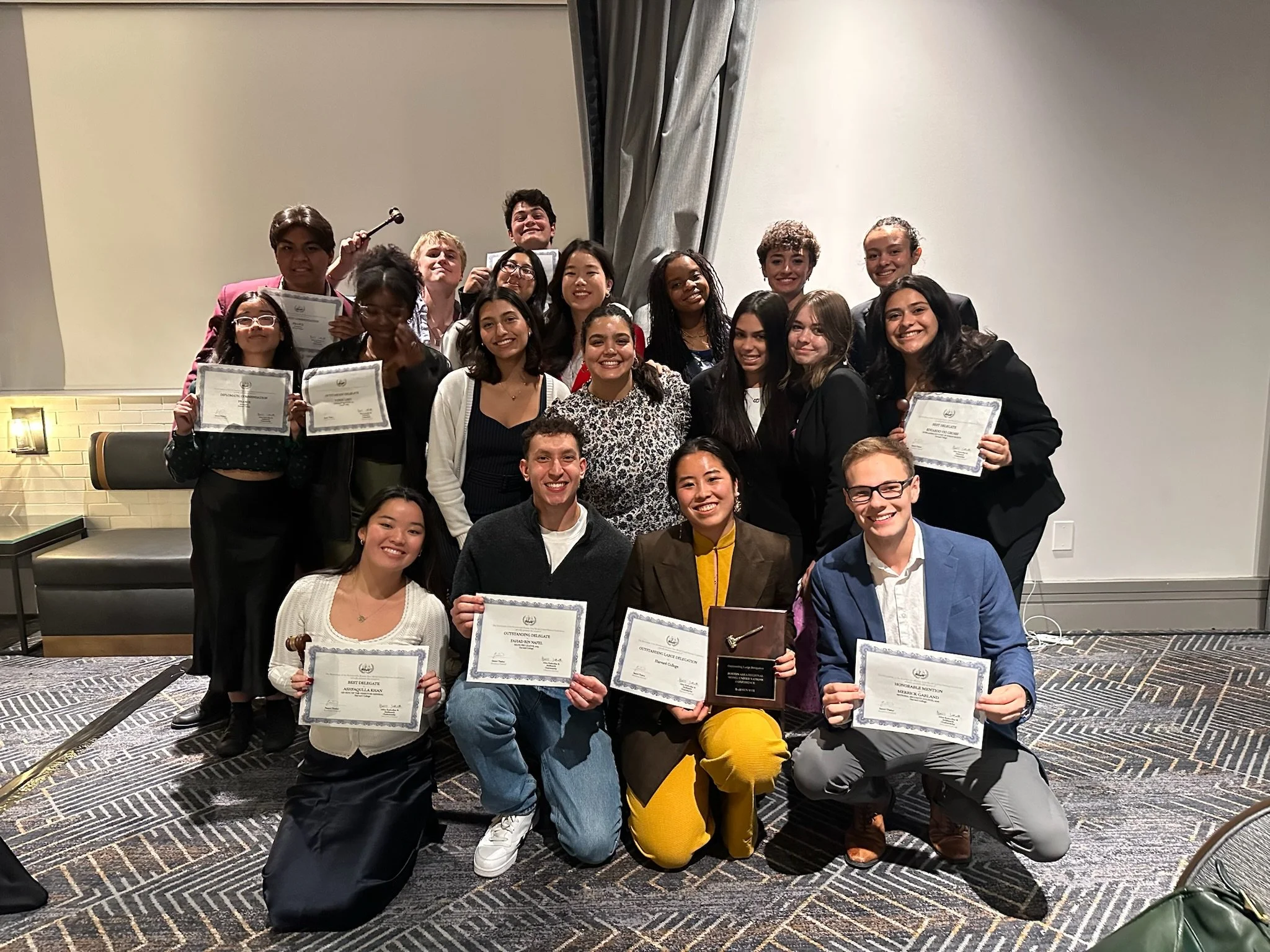 A group of young people and adults posing together in a conference room, holding certificates and trophies, celebrating their achievements at an award ceremony.