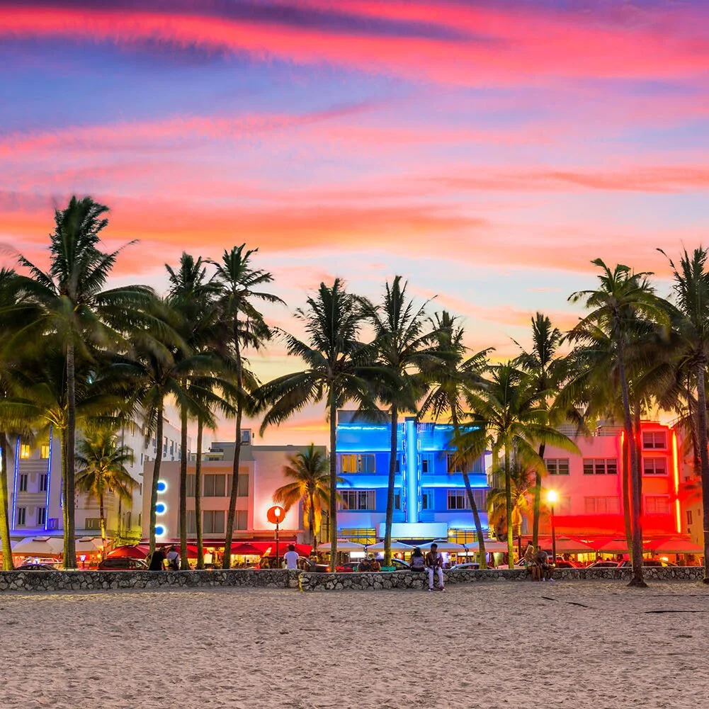 Colorful sunset sky over a beach with palm trees and neon-lit buildings.