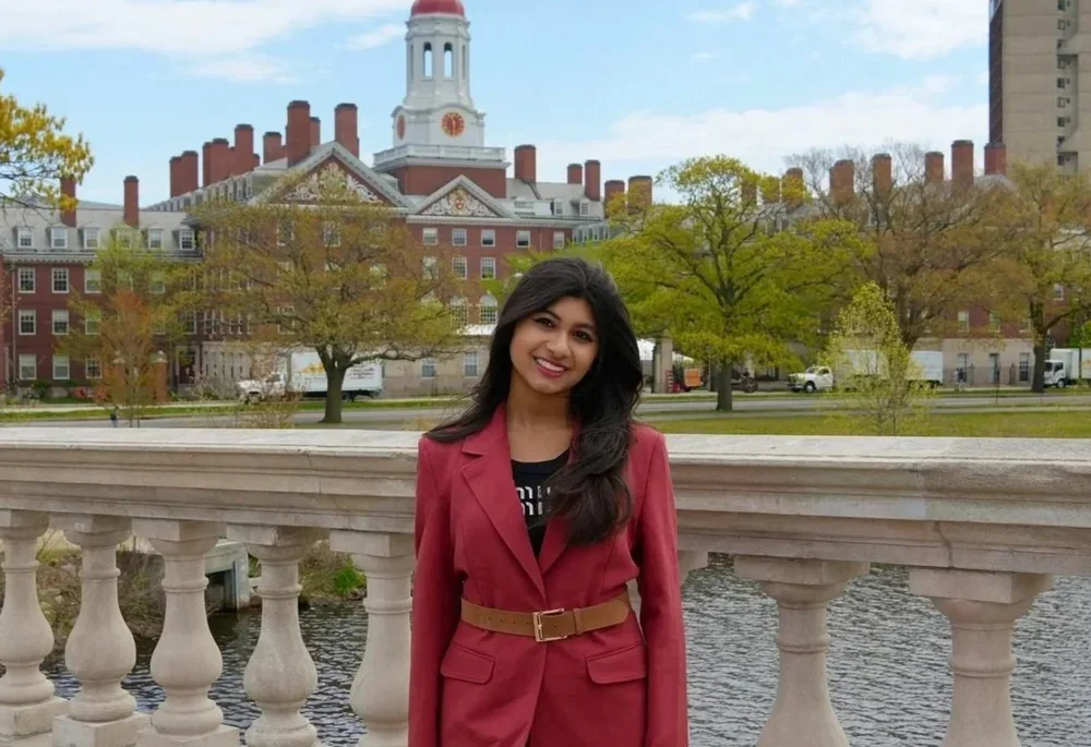 A woman with long dark hair, wearing a red blazer and belt, smiling, standing on a stone bridge with a historic building and trees in the background.