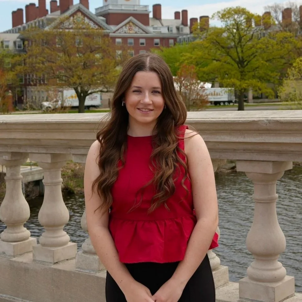A young woman with long, wavy brown hair wearing a sleeveless red top and black pants, standing on a bridge with stone balustrades, smiling at the camera. Behind her, there are trees with green and yellow leaves, a river, and a backdrop of buildings with red brick facades and multiple chimneys.