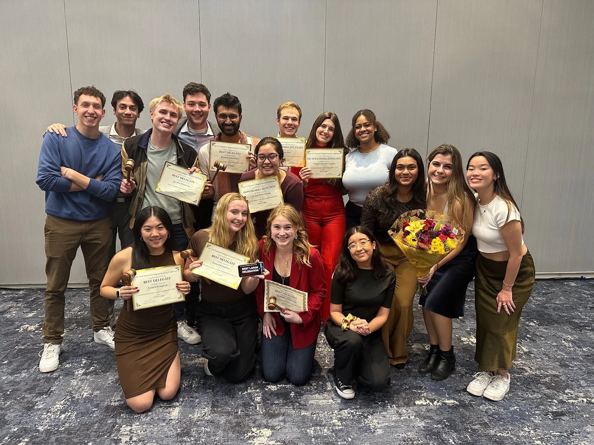 Group of young adults at an award ceremony holding certificates and flowers, standing in front of plain wall.
