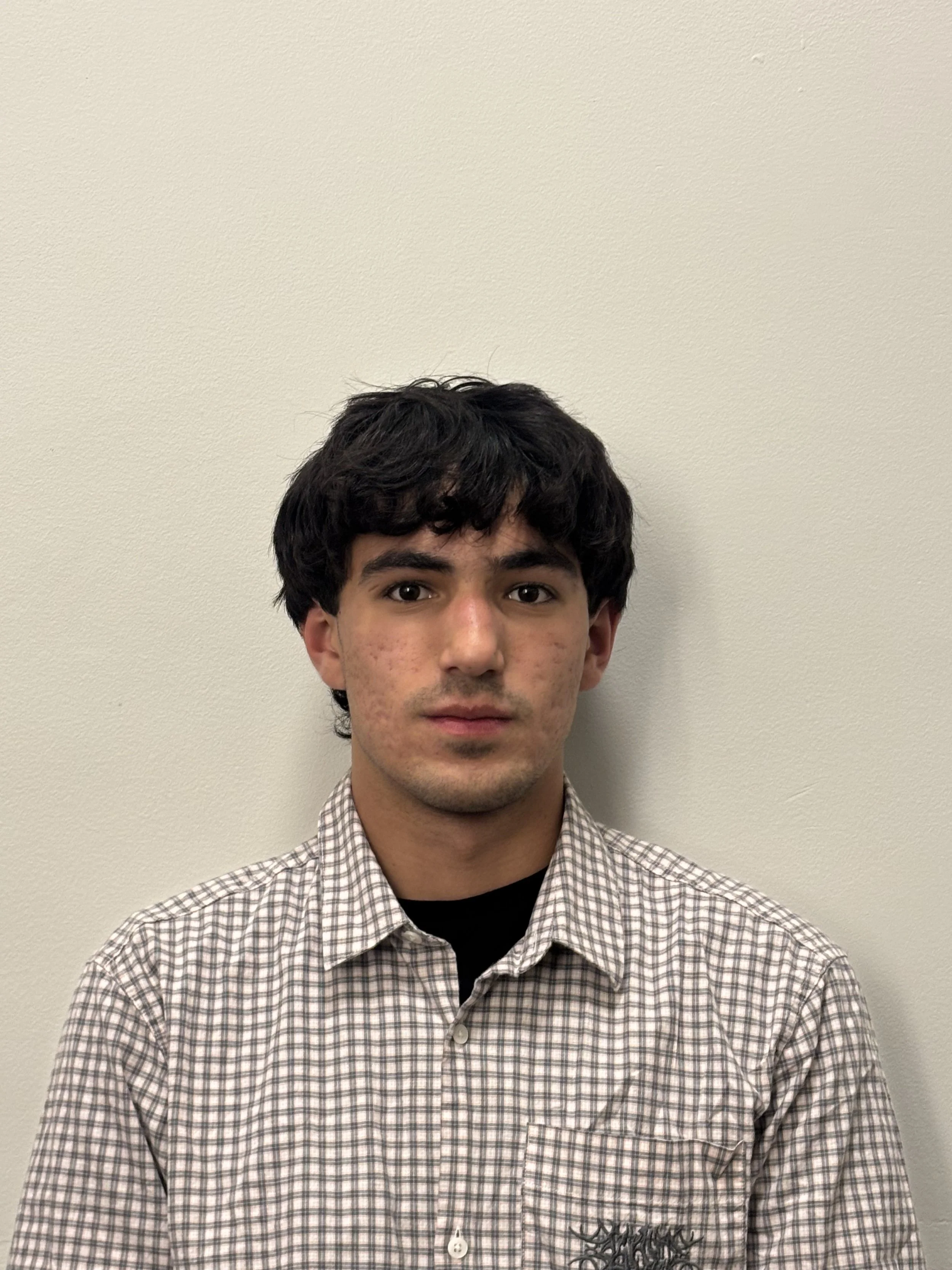 A young man with dark, curly hair and light skin, wearing a plaid shirt over a black shirt, stands against a plain beige wall.