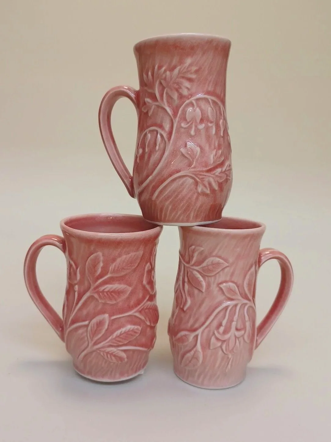 Three pink ceramic mugs with carved floral and leaf patterns, arranged with one mug on top of two others against a plain background.