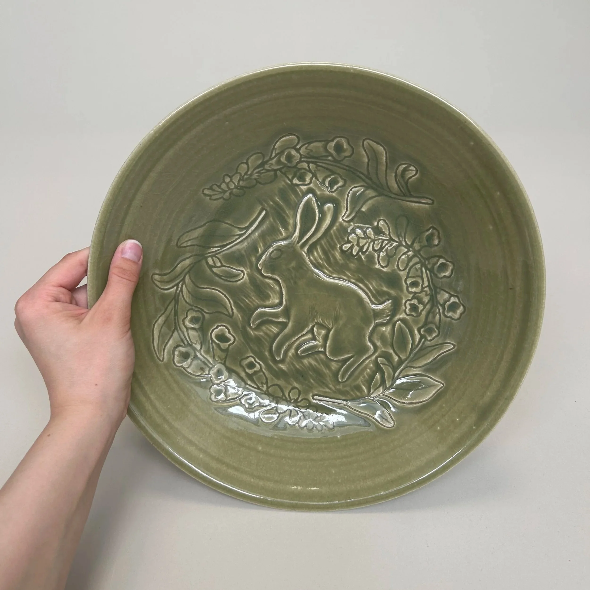 A person holding a green ceramic plate with a raised floral design, against a plain light background.