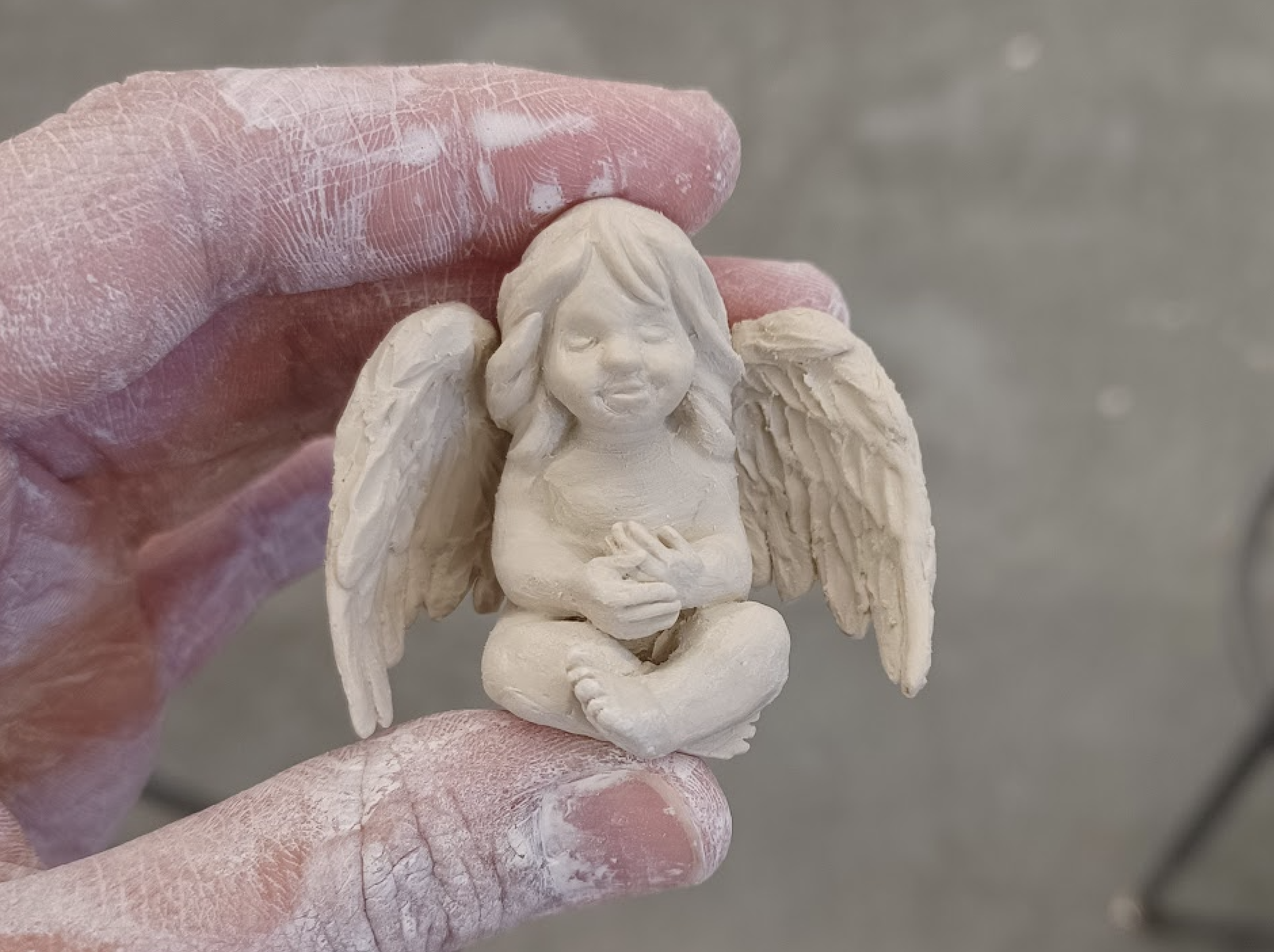 A sculptor's hand holding a small, detailed clay statue of an angel, sitting cross-legged with a peaceful expression.
