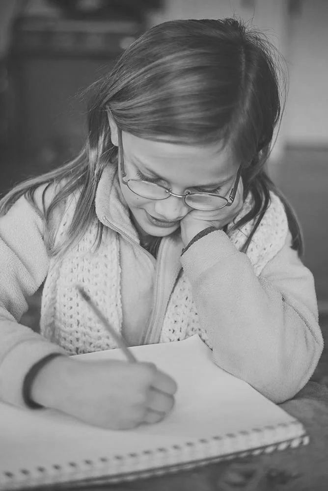 A girl with glasses is sitting at a table, drawing in a notebook, and resting her head on her hand.
