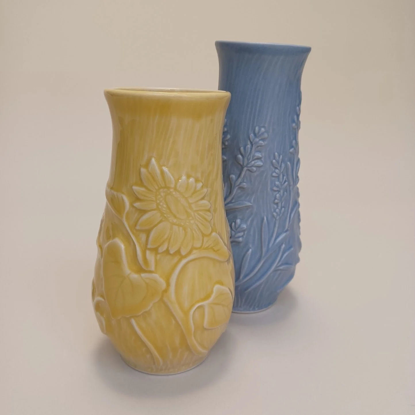 Two ceramic vases, one yellow and one blue with floral patterns, positioned on a white surface against a plain beige background.