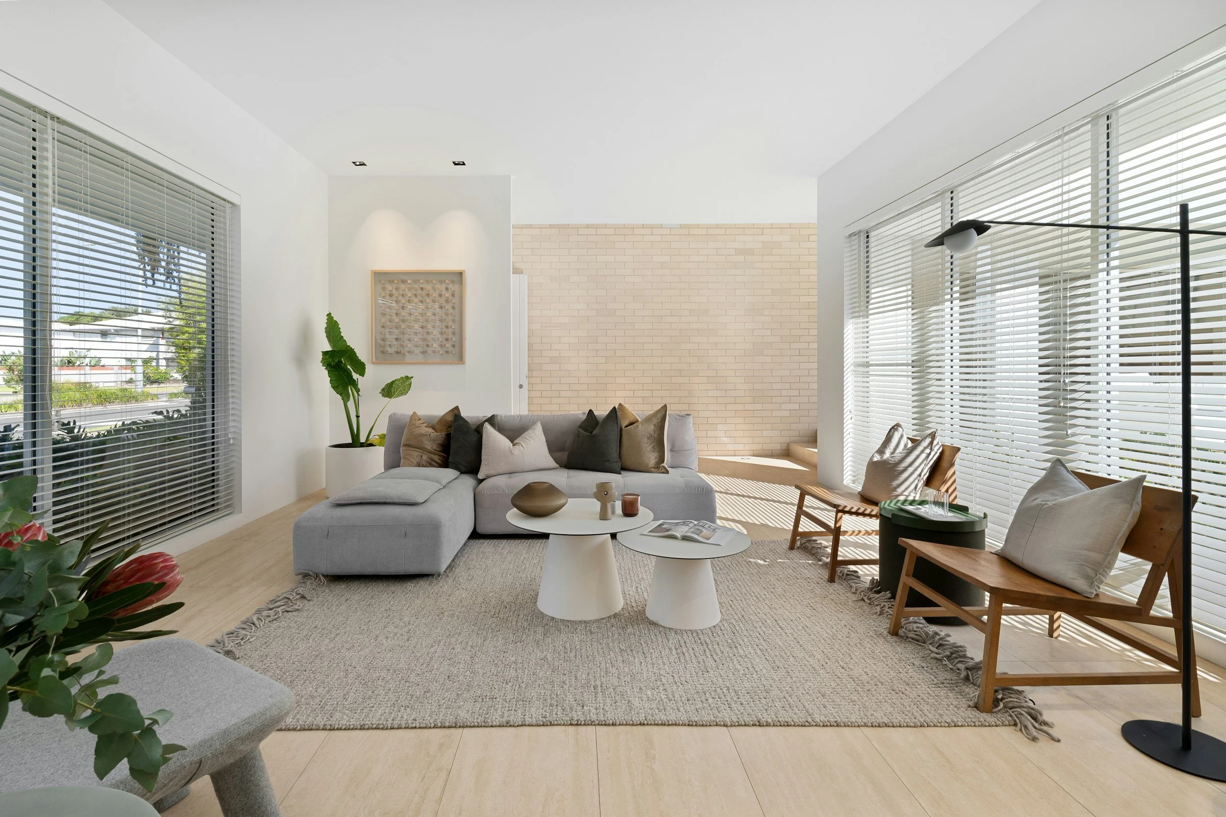 Bright living room with large windows with white blinds, light-colored walls, and a beige brick accent wall. Contains a gray sectional sofa with multiple cushions, two white round coffee tables, and potted plants. A black floor lamp with a modern design is also visible.