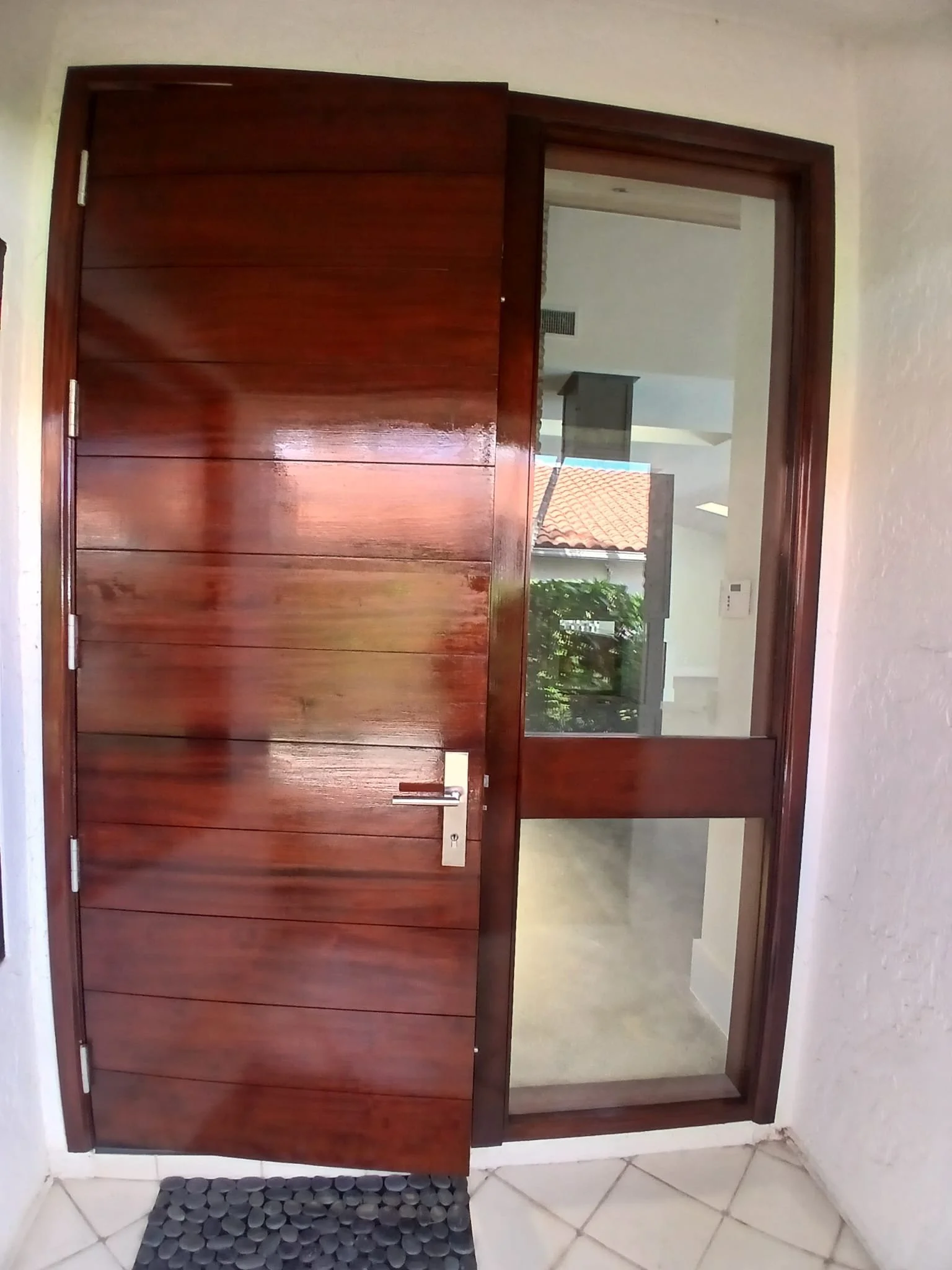 A wooden front door with a glass window, set in a white wall. The door is closed, and the area outside shows a glimpse of a roof with red tiles and some greenery. There is a black textured doormat in front of the door on beige tiled flooring.