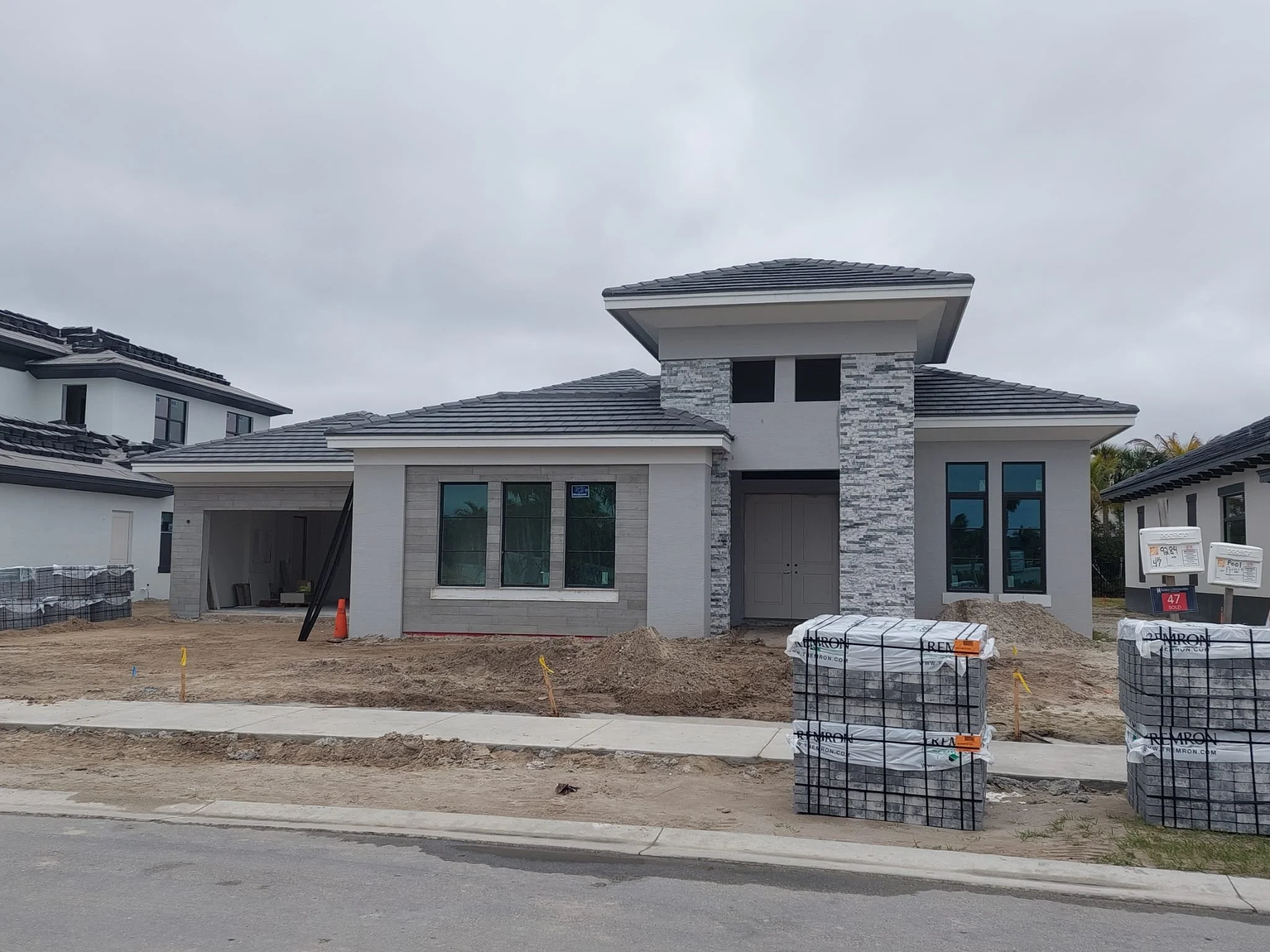 A modern house under construction with a gray shingled roof, a stone facade, and large windows. Construction materials and supplies are visible in the front yard, with cloudy skies above.