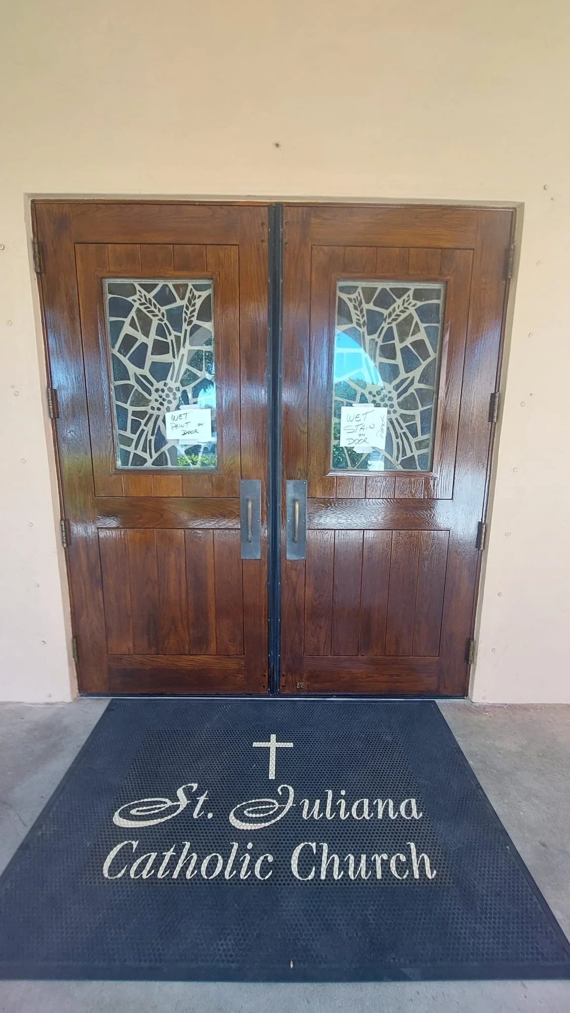 Double wooden doors with decorative glass panels, signs with wet stand instructions, and a black mat with the St. Juliana Catholic Church logo.