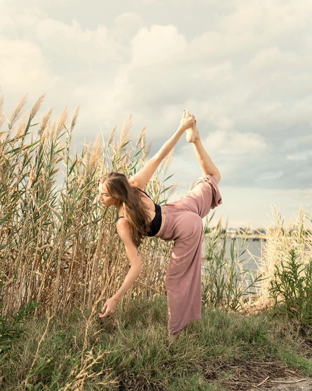 Claire Bilavsky in naṭarājāsana (dancer pose) among tall reeds beside the water.
