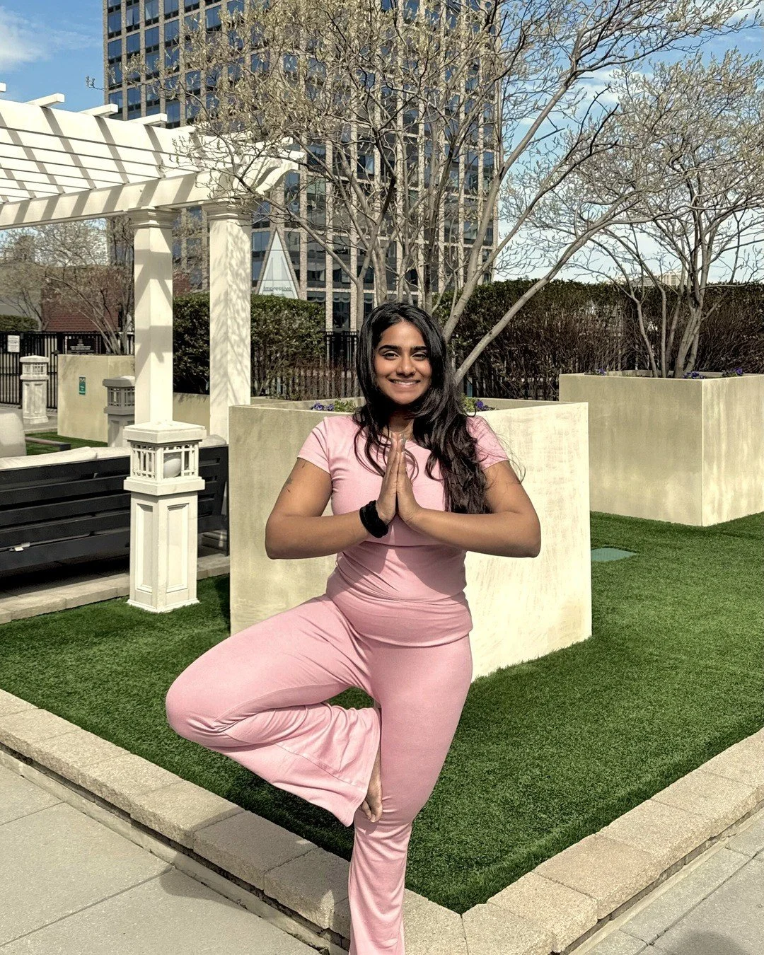 Nandita Shyam in vṛkṣāsana (tree pose) with hands at her heart center in añjali mudrā, standing on a lawn in a courtyard.
