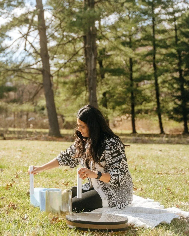 Seema Vachhani, sound healing practitioner at ĀTMĀ Yoga Collective, kneeling on a blanket in a sunlit field with her crystal singing bowls.