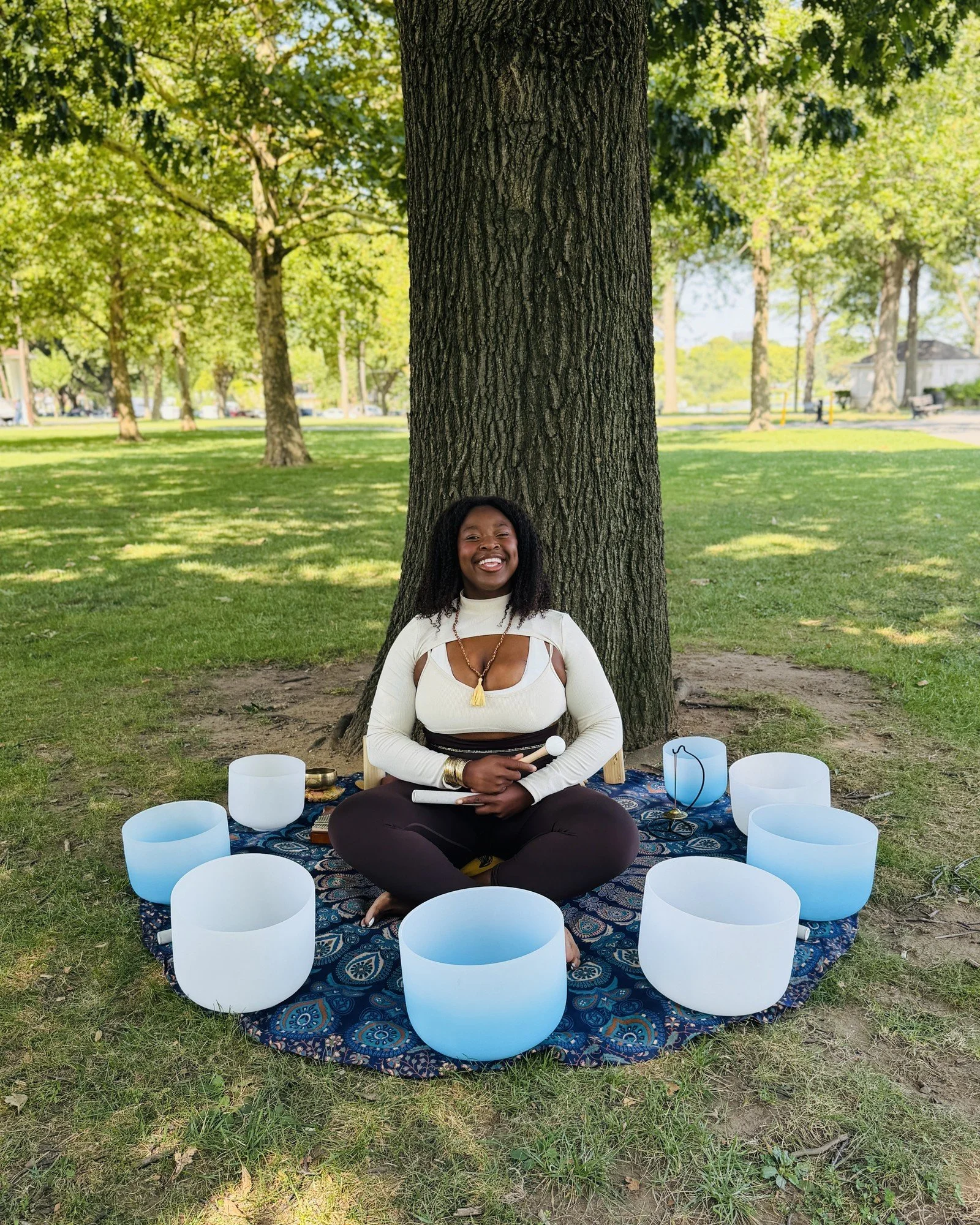 Baldween Casseus seated among a ring of crystal singing bowls on a patterned cloth, beneath a large tree.