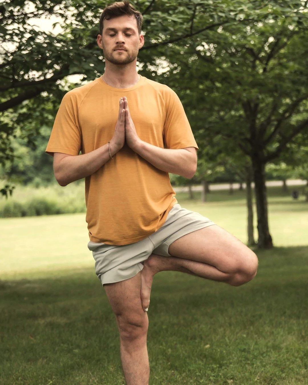 handler Lennon standing in vṛkṣāsana (tree pose) on the grass in a leafy park, eyes closed, hands in añjali mudrā (palms together) at his chest.