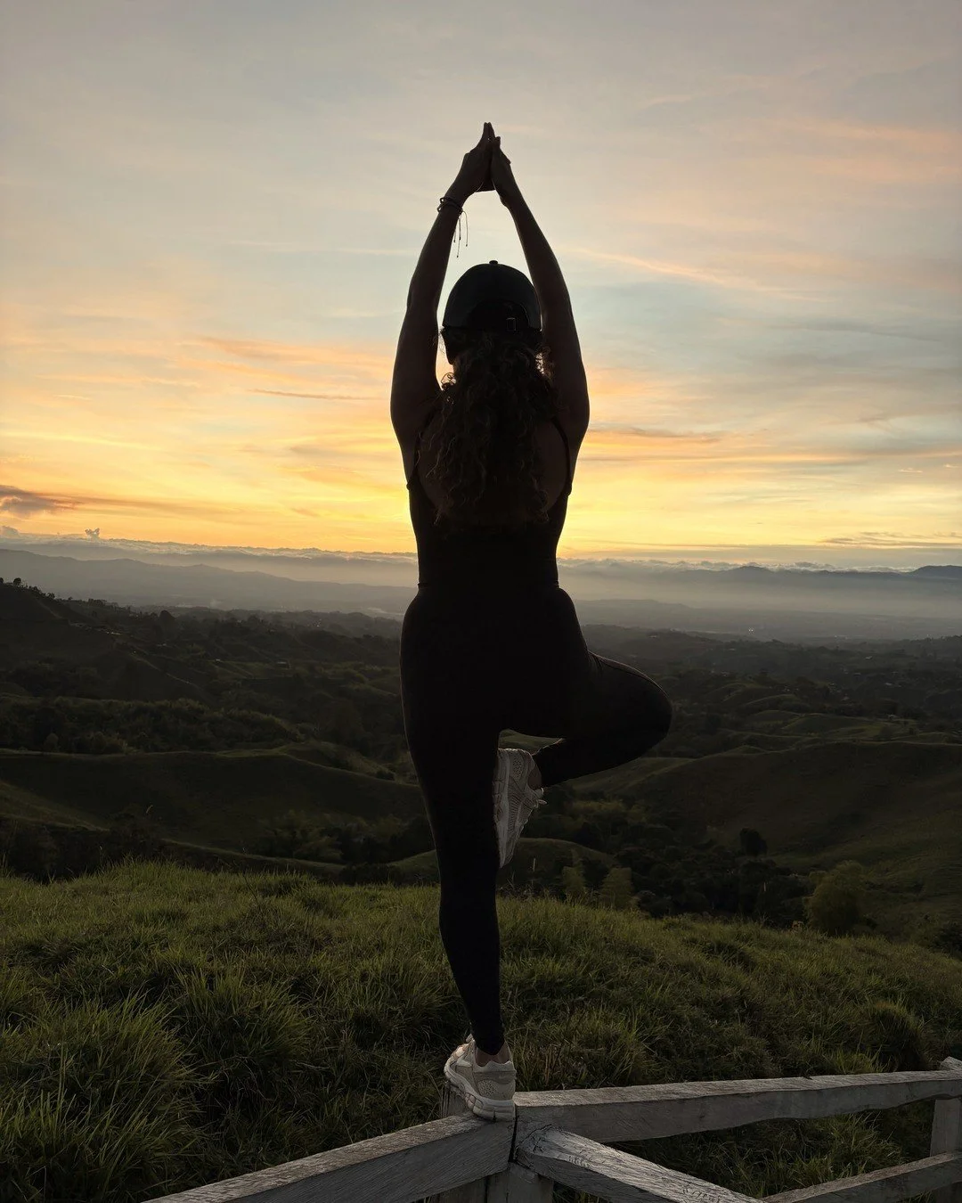Angelica Rivera in vṛkṣāsana (tree pose), in silhouette against a sunrise over open hills, arms overhead and hands in kālīmudrā.