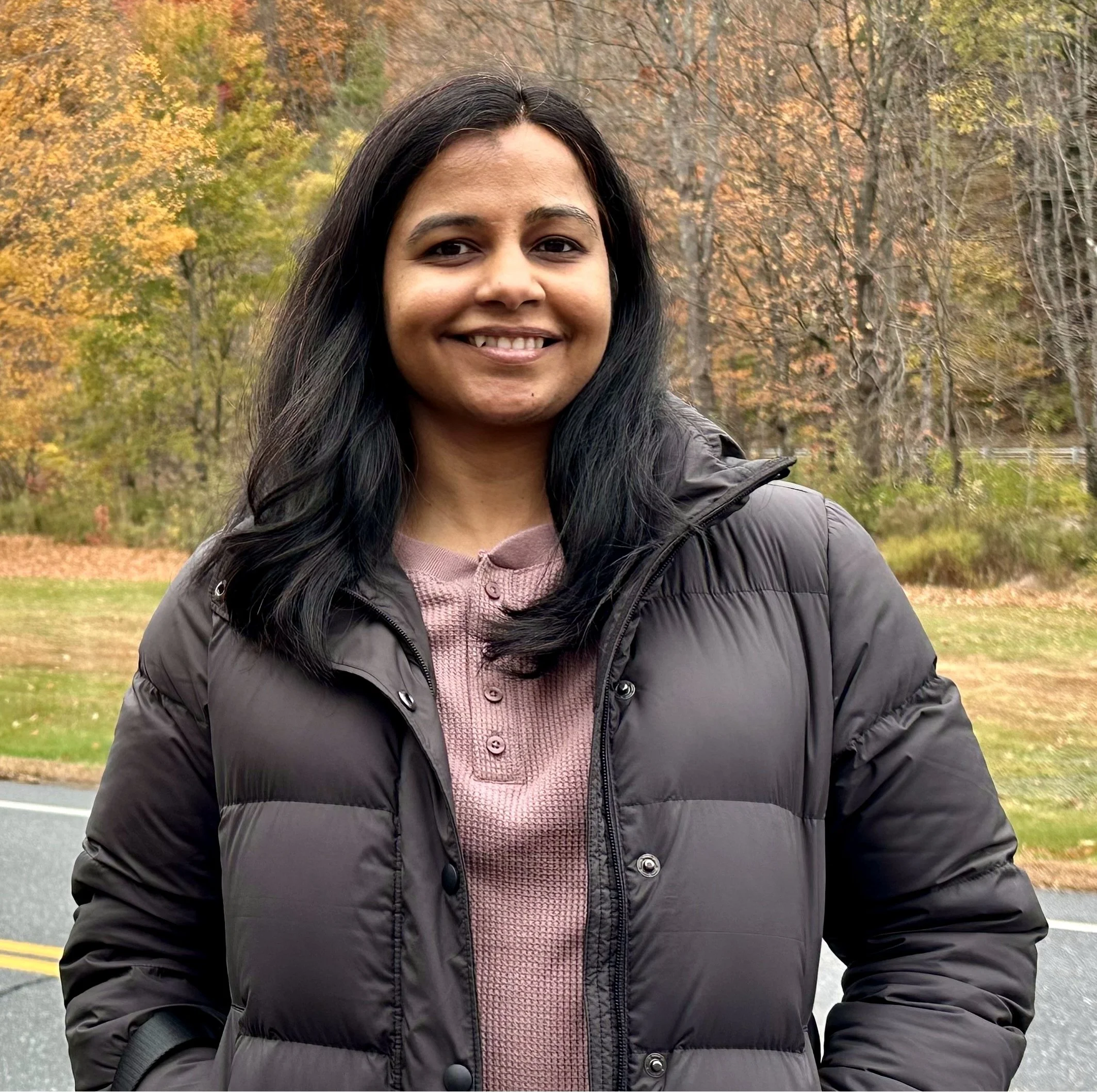 Anumeha Taori, Vinyāsa & Prāṇāyāma teacher at ĀTMĀ Yoga Collective, smiling outdoors among autumn trees.