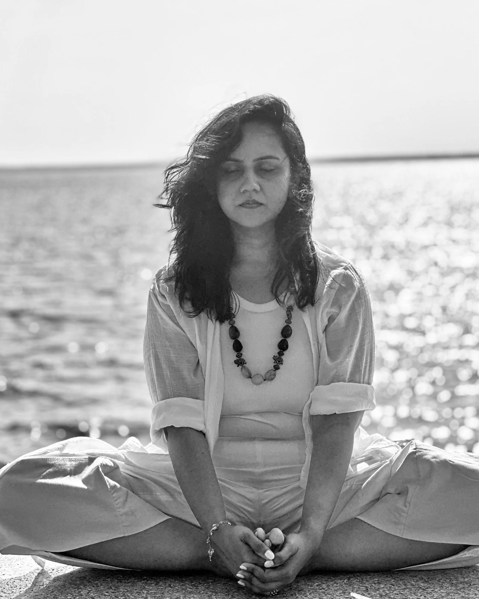 Monisa Parekh seated in baddha koṇāsana (seated butterfly) beside open water with her eyes closed, photographed in black and white.