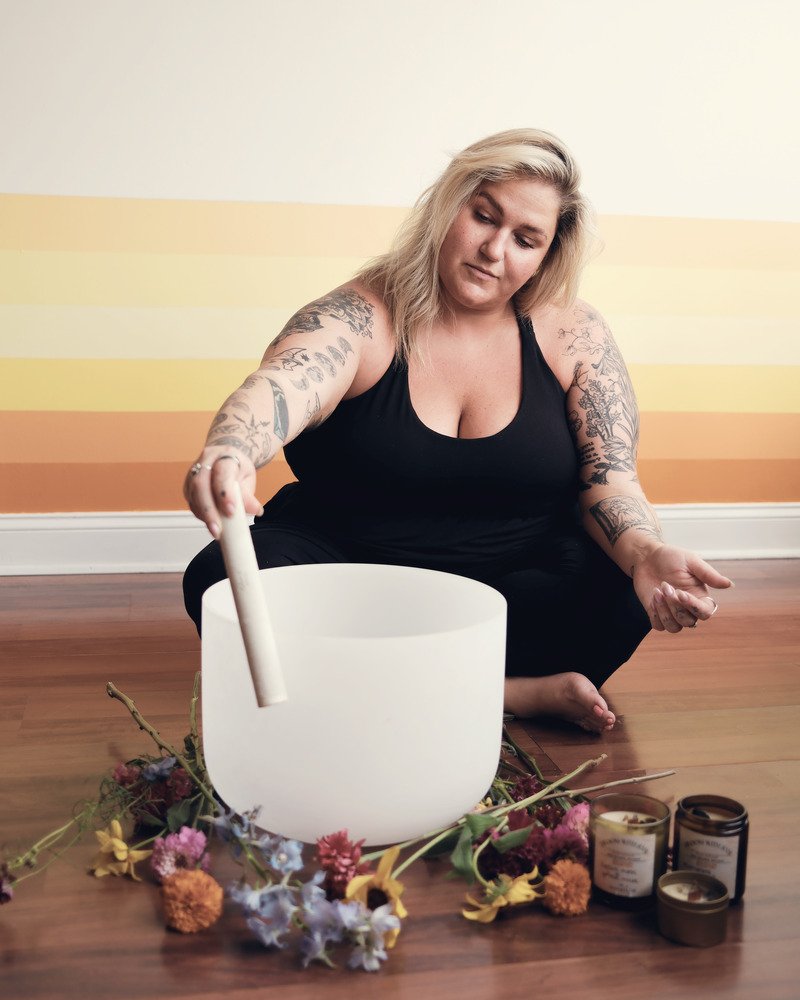 Sam, a reiki and yoga teacher, sits cross-legged on a wooden floor beside a white crystal singing bowl, holding its mallet, with dried flowers and small candles arranged in front of her.